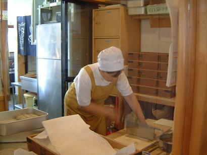 白い割烹着に黄色いエプロンと白帽子をかぶった女性が、木枠の箱の上に置かれた生地を丁寧にそばを切る様子の写真