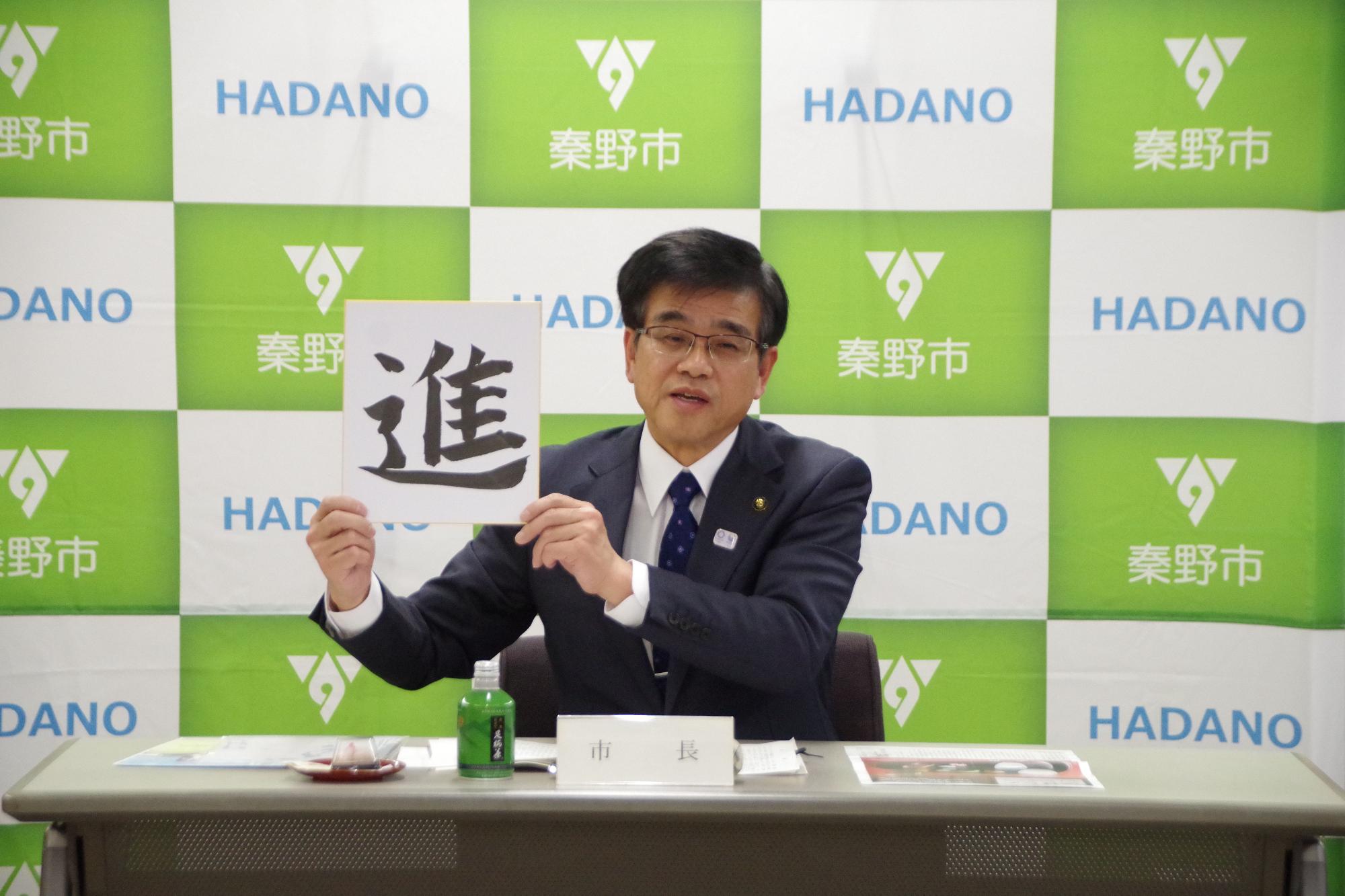 秦野市のバックパネルの前で、札や飲み物のペットボトルが置かれた机に座ったスーツ姿の市長が、「進」と大きく書かれたボードを掲げている、1月記者会見の様子の写真