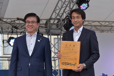 たばこ祭会場の白いテントの下で、スーツ姿の市長と木材で作られた任命書を持っている吉田栄作さんが並んで立っている授与式の様子の写真