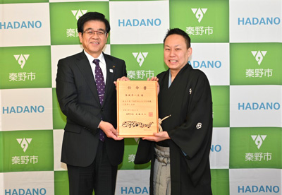 黄緑と白の秦野市のバックパネルの前で、スーツ姿の市長と黒い紋付の着物と袴を着用した春風亭一左さんが木材で作られた任命書を一緒に持ち立っている授与式の様子の写真