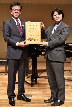 グランドピアノの前で、スーツ姿の市長と山田和樹さんが木材で作られた任命書を一緒に持ち立っている授与式の様子の写真