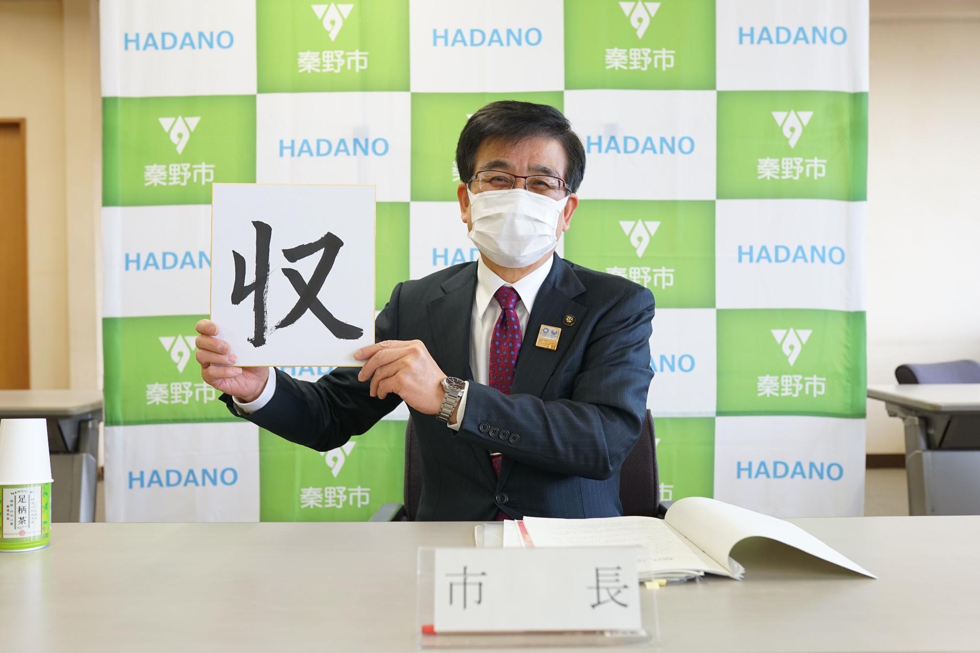 秦野市のバックパネルの前で、「市長」と書かれた札と資料が置かれた机に座り、マスクをつけたスーツ姿の市長が、「収」と大きく書かれた色紙を掲げている、1月定例市長記者会見の様子の写真