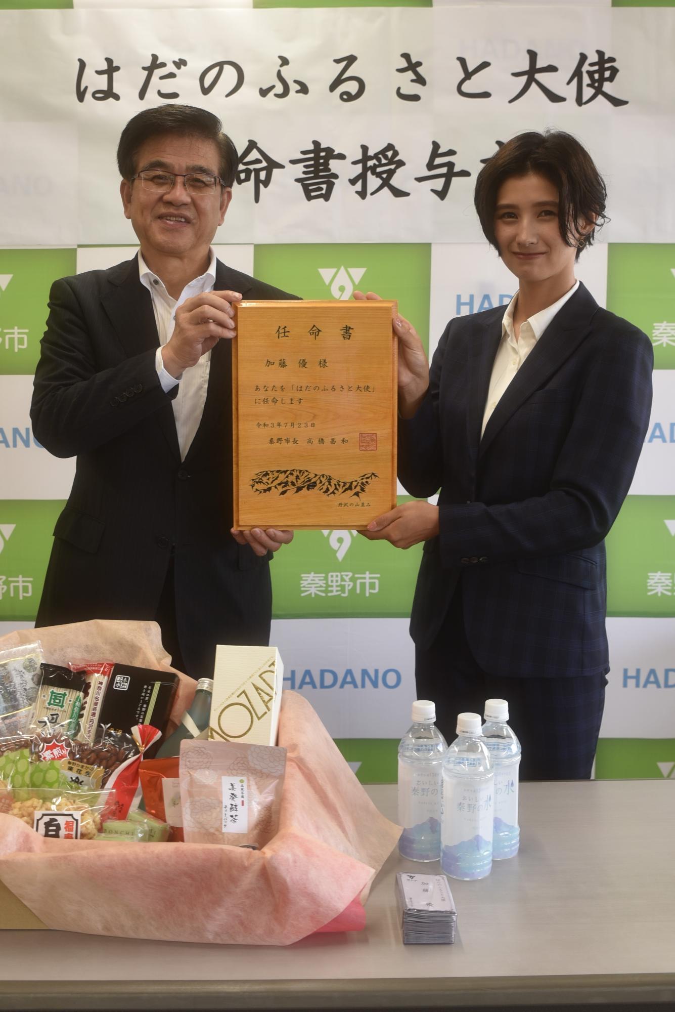 「はだのふるさと大使任命授与式」と書かれた秦野市のバックパネルと、沢山の特産品や水が入ったペットボトル、名刺が置かれた長机の前で、スーツ姿の市長と加藤優さんが一緒に木製の任命書を持ち並んでいる任命書授与式の様子の写真