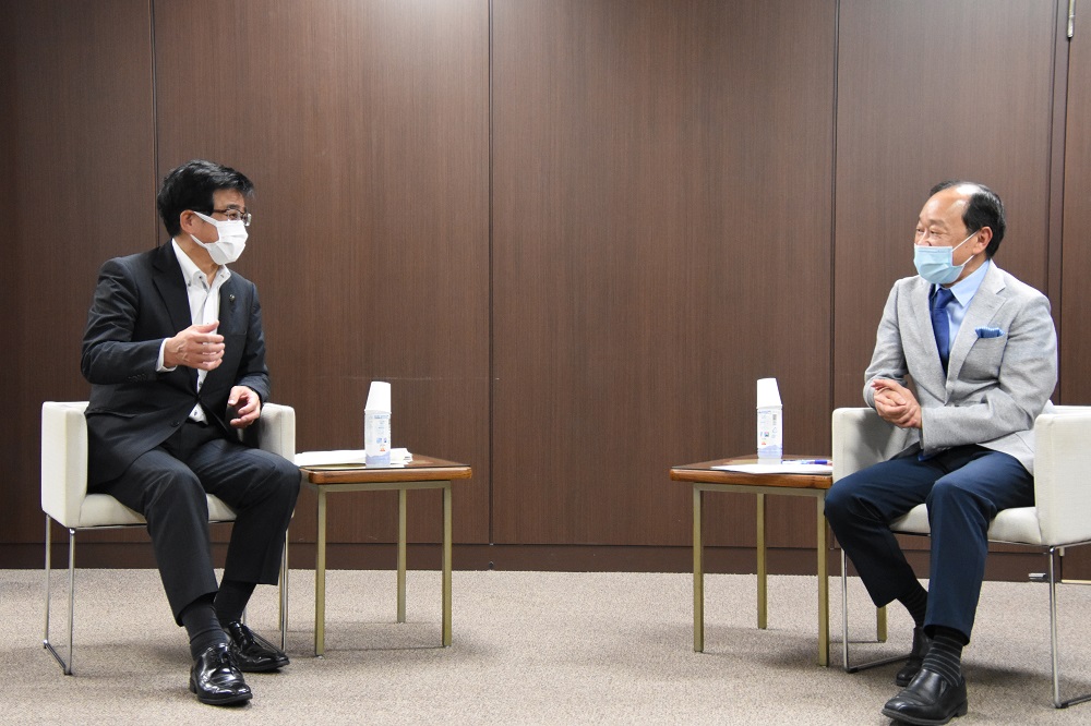 茶色い木目調の壁の部屋で、マスクを着用した二人のスーツ姿の男性が、それぞれ小さなサイドテーブルか置かれ間隔を空けて白い一人掛けの椅子に座り対談している様子の写真