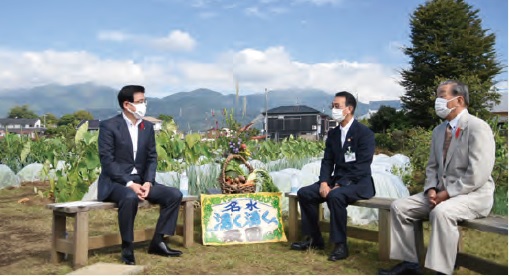 遠景には山々が広がり、中央には「名水湧く湧く農園」と書かれたボードと収穫物が入った籠が置かれた広大な畑の真ん中で、スーツ姿の三人の男性がベンチに座って対談している様子の写真