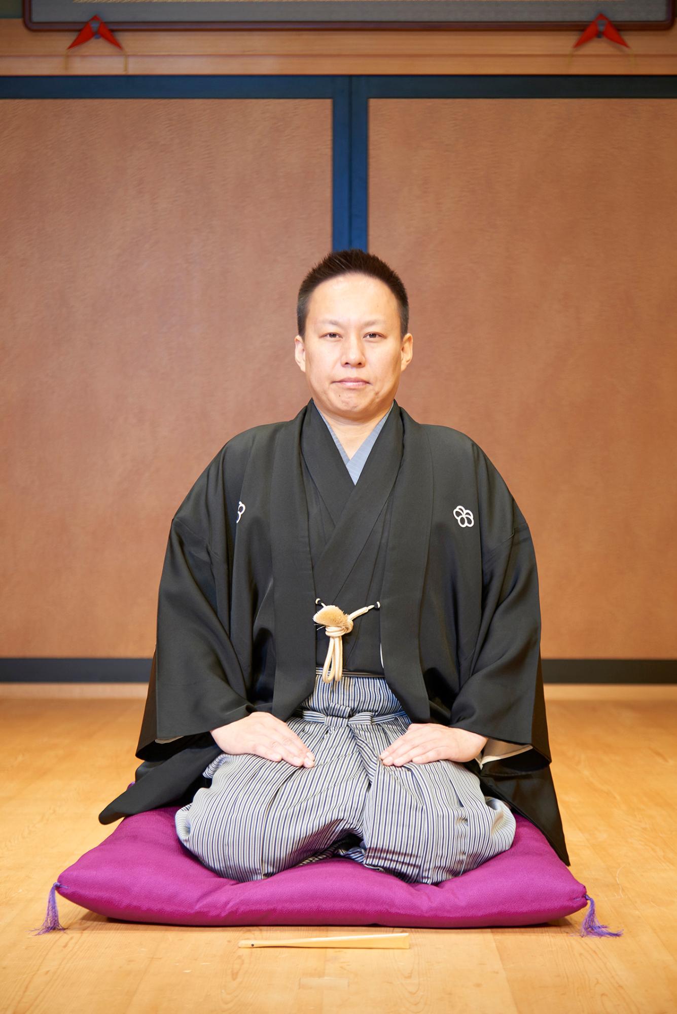 木目調の床と襖のような背景の部屋で、黒い紋付の着物と袴を着用した春風亭一左さんが、紫色の座布団の上に正座しており、正面を向いて静かに座っている写真