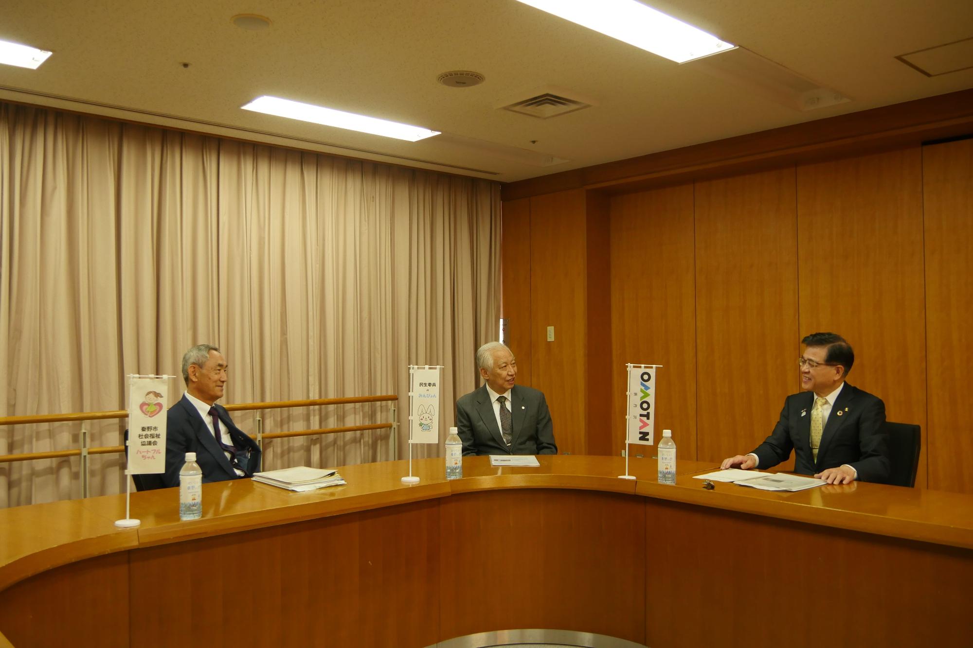 木目調の壁と薄いベージュのカーテンの会議室で、資料やペットボトルが置かれ、旗が立てられているコの字型の大きなテーブルに三人のスーツ姿の男性が間隔をあけて座っている写真