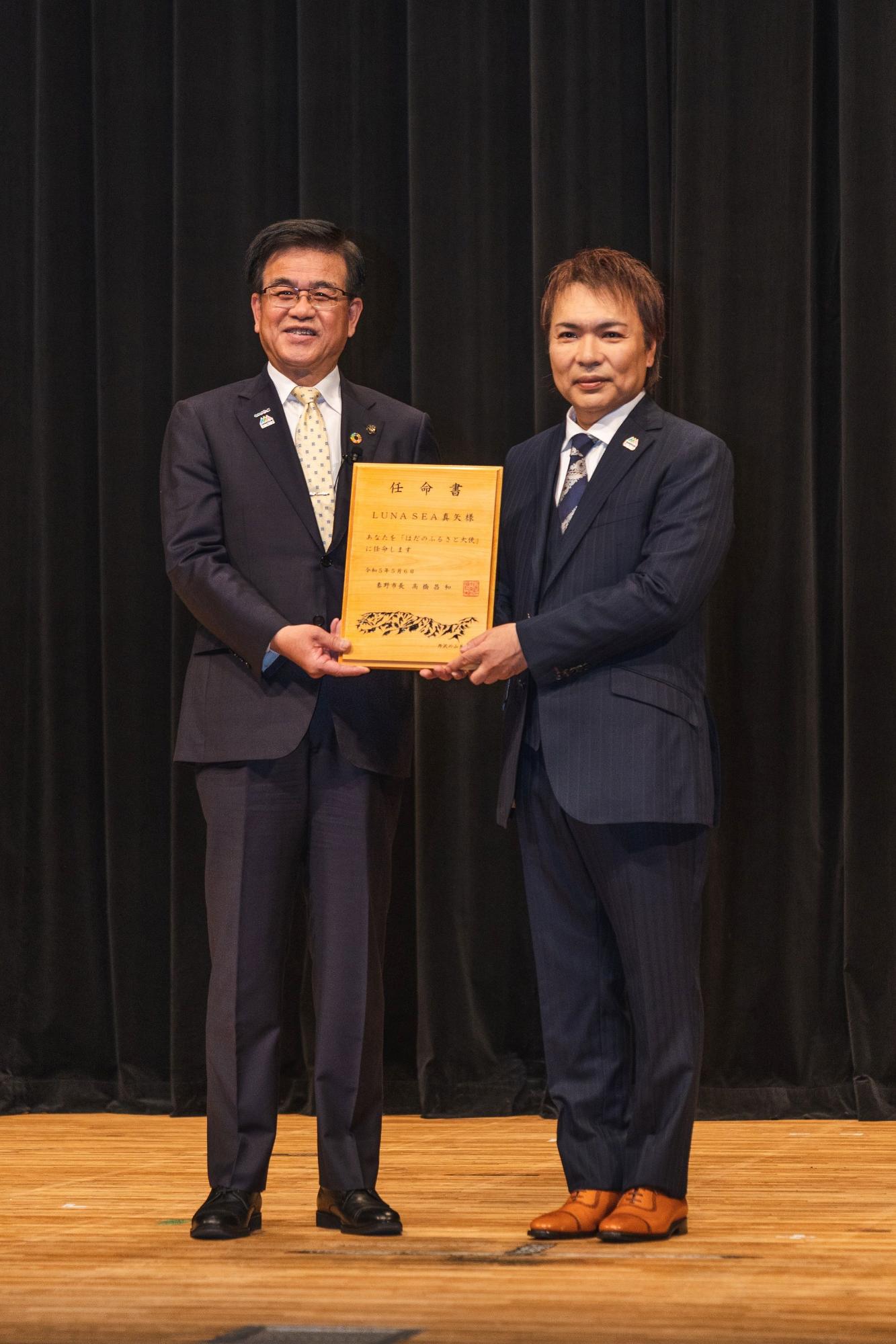 黒い幕の前で、スーツ姿の市長と真矢さんが木材で作られた任命書を持ち並んで立っている授与式の様子の写真