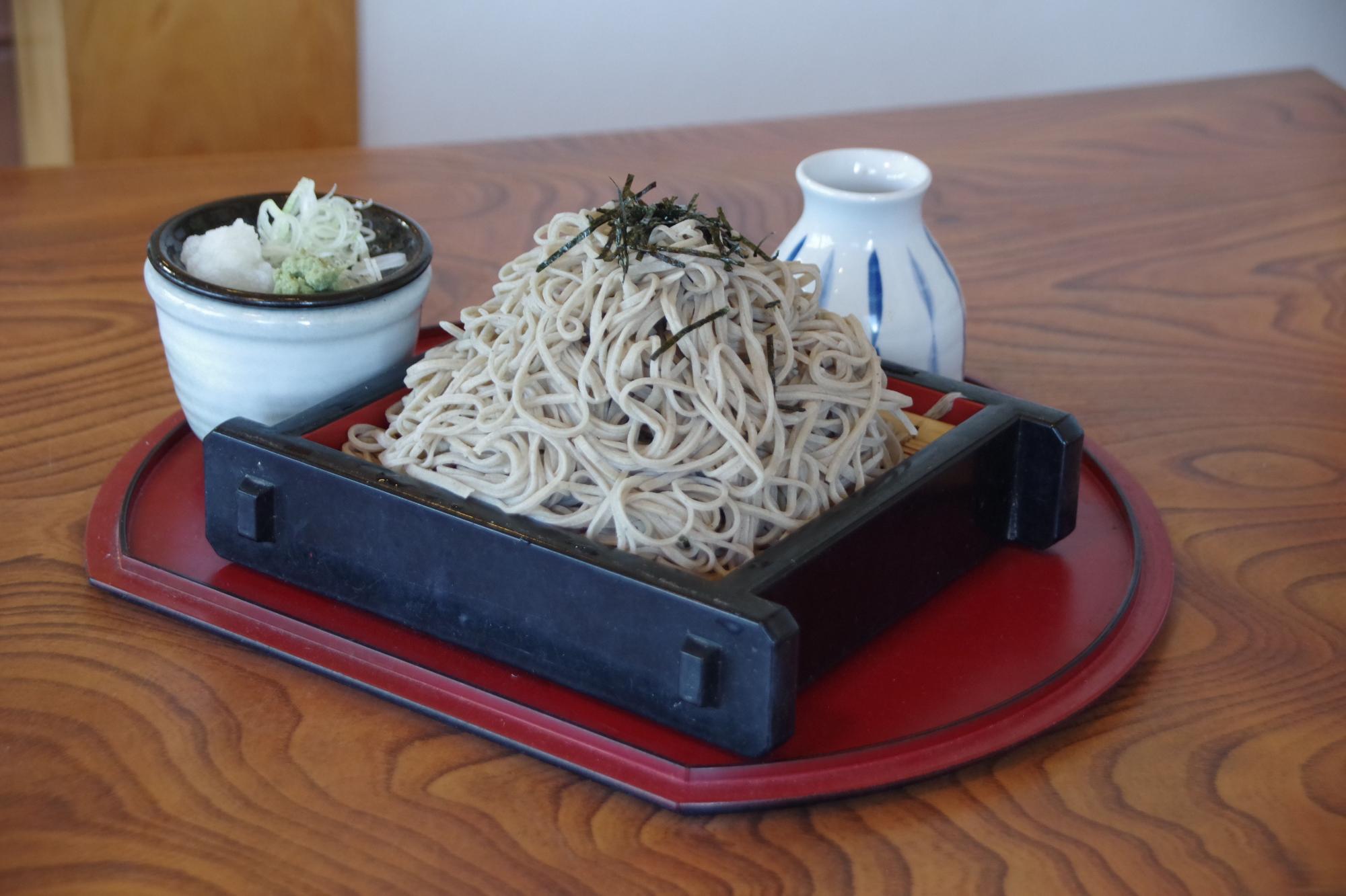 木目調のテーブルの上で、赤いお盆に乗せられたざる蕎麦と、つゆの入った徳利、薬味の入ったそば猪口が揃っている様子の写真