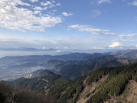 雪をかぶった富士山が右奥に見え、手前には針葉樹の森が広がる急峻な山並みが幾重にも重なり、その遠景には広がる霞が望める、晴れた日の山頂からの雄大な風景の写真