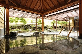 開放感のある木造の屋根と柱に囲まれた空間に、石に囲まれた湯船が配置されており、湯気が立つ静かな水面には周囲の景色が映り込み、竹垣で囲まれた奥には緑の植栽が配置された、露天風呂の室内風景を捉えた写真