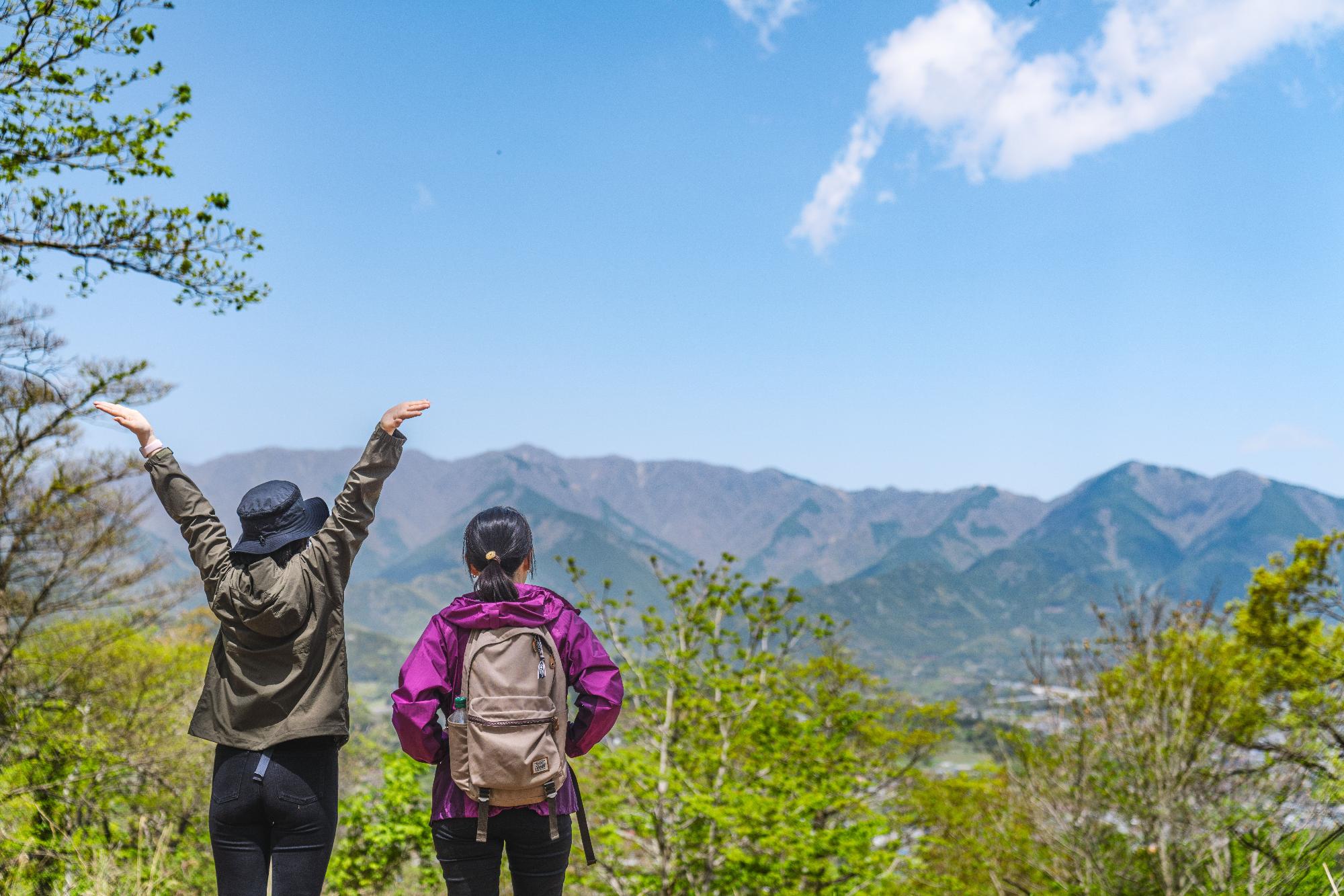 ハイキング中の2人の女性が山の頂上から壮大な景色を眺めており、1人は帽子をかぶって両手を広げている後ろ姿の写真