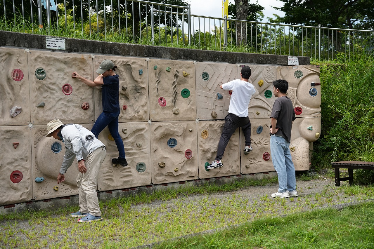 屋外の公園にある高さの低いボルダリング用の壁で、年代の違う男性たちがそれぞれ壁を登ったり、登る仲間を眺めたりして楽しんでいる写真