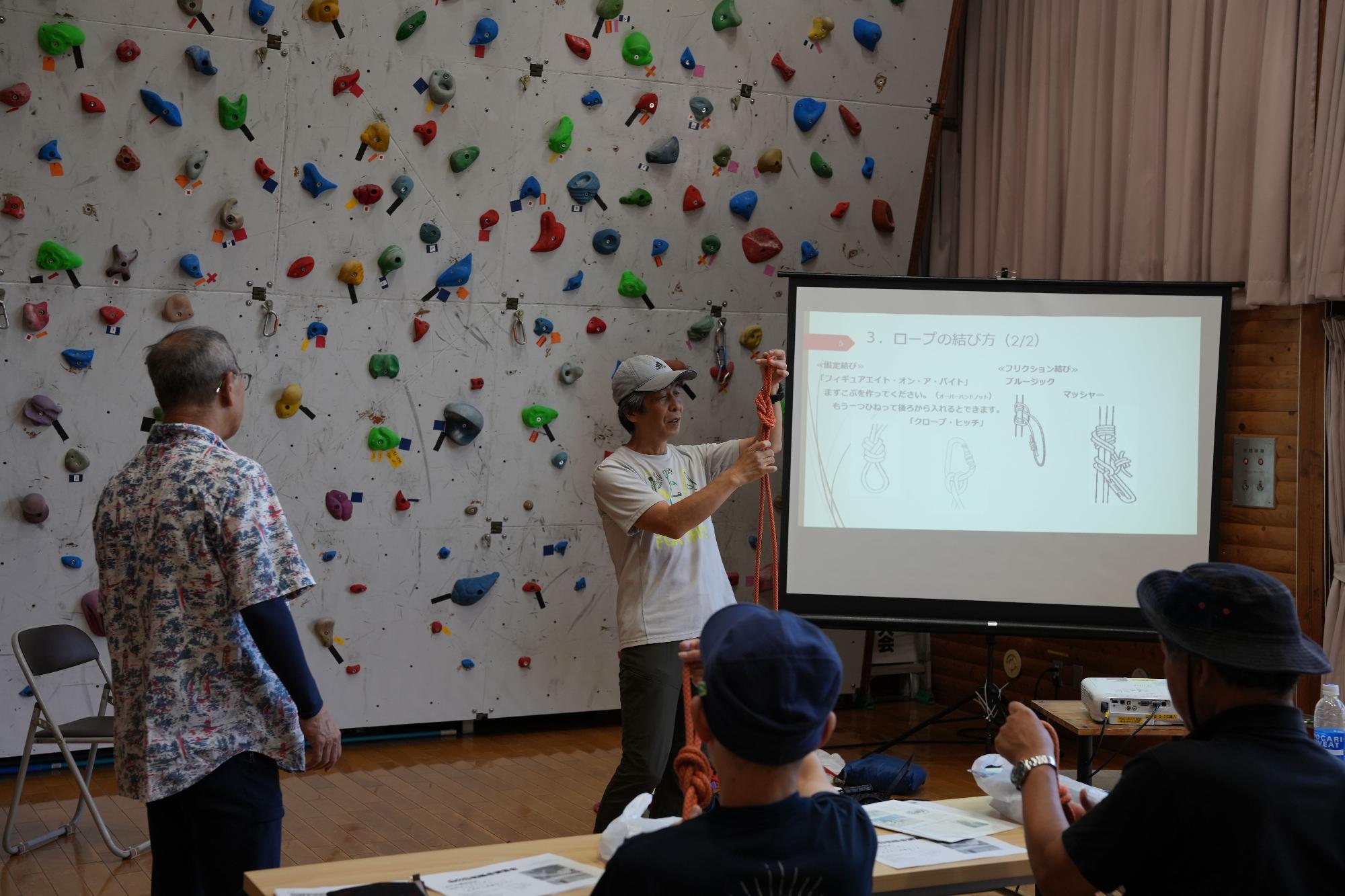 クライミングウォールがある室内で、講師がロープの結び方を実演し、スクリーンには結び方の図が表示されている講習会の様子の写真