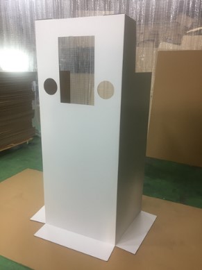 緑色の床の上に立てられた、中央に透明な窓と両側に円形の穴が開いた白い段ボール製の検査用防御フェンスの写真