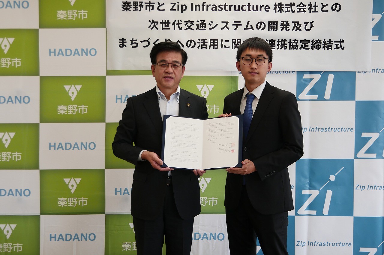 秦野市とZip Infrastructure株式会社のバックパネルを背景に、市長と須知代表取締役が「秦野市とZip Infrastructure株式会社との次世代交通システムの開発及びまちづくりへの活用に関する連携協定締結式」と書かれたボードの下で、開いた協定書を両手で持ち、カメラに向かって立っている写真
