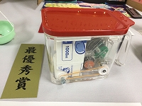 「最優秀賞」と書かれた金色の短冊が置かれており、赤い蓋がついた透明のプラスチック容器の中に牛乳パックやモーター、輪ゴム、などが組み込まれた手作りの作品が展示されている写真