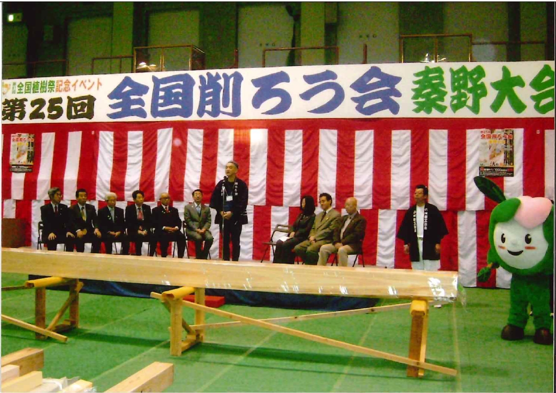 広い屋内会場で、「全国削ろう会 秦野大会」と書かれた横断幕の下、正装した関係者が雛壇に座り、中央の男性がマイクで話し、手前には長い角材が置かれ、右端には緑色のマスコットキャラクターが立っている写真