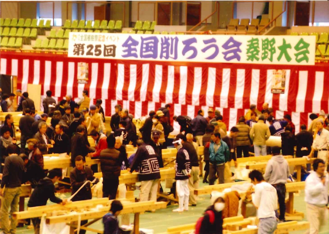 「第25回 全国削ろう会 秦野大会」と書かれた横断幕の下、赤白の幕が張られた中央奥のステージ手前に大勢の参加者が集まり、作業台らしき木製のテーブルが並ぶ広いフロアで削る作業や交流を楽しんでいる写真