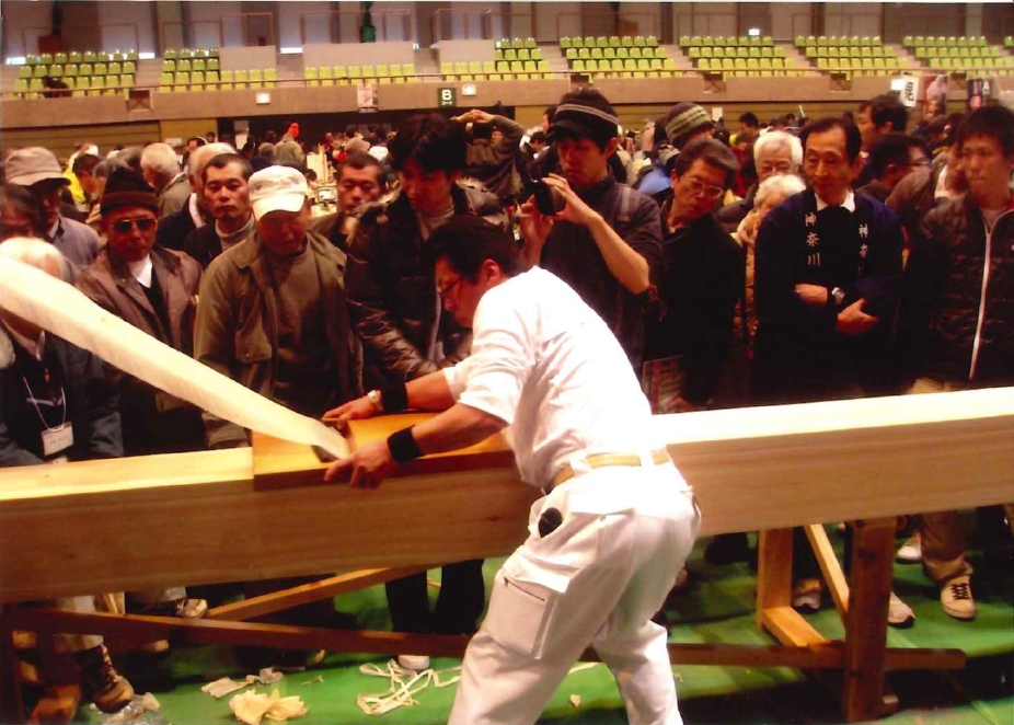 真剣な面持ちの男性が、多くの観客に取り囲まれた会場で、目の前の木製の作業台に固定された太い角材に力を込めてかんなをかけ、長く薄く白い削り華を生み出している瞬間を捉えた写真