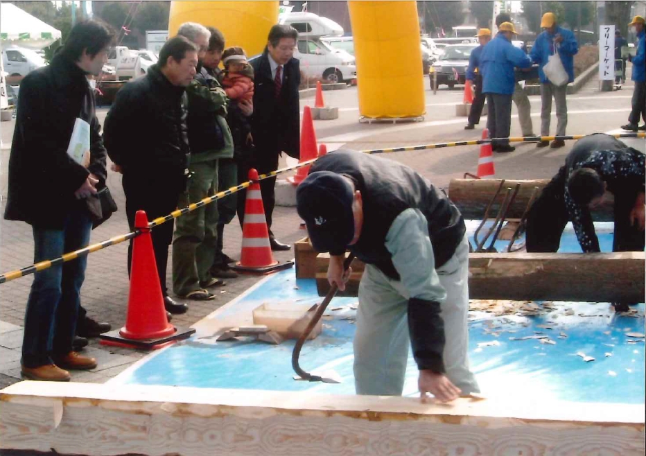屋外イベント会場で、キャップを被った男性が土台に固定された大きな丸太に向かって曲がった刃のついた道具を振り下ろし、作業を見守る数人の見物客の背景には、黄色いアーチや三角コーンが見える作業風景の写真
