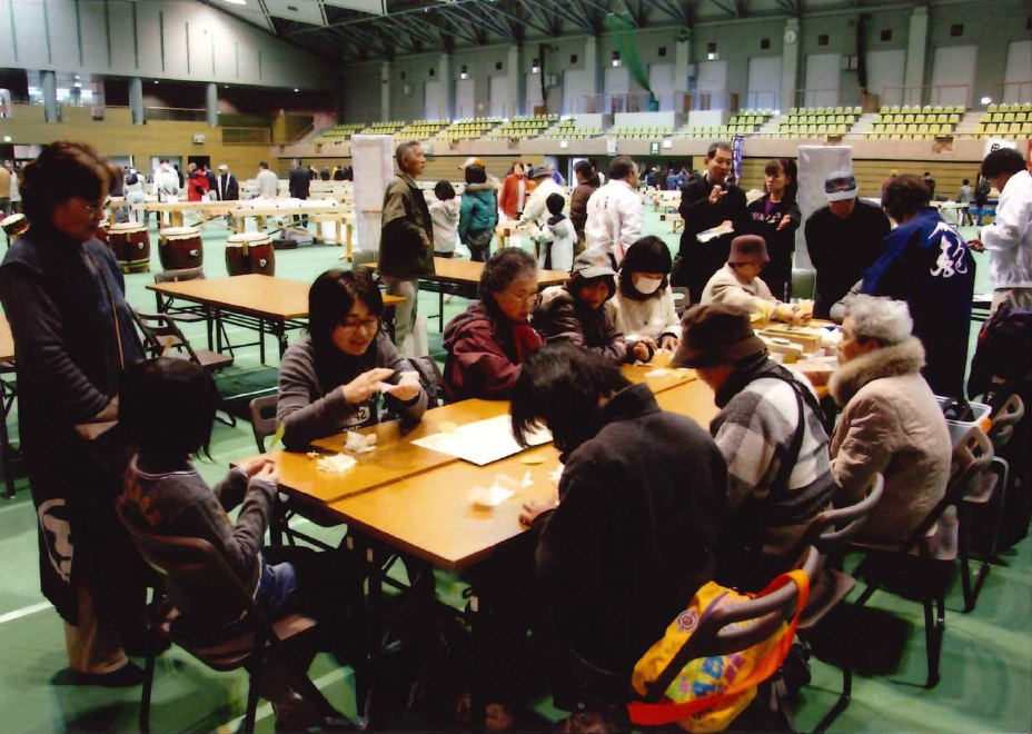 広い体育館のような屋内会場で、複数の参加者が長机に集まって休憩や談笑をしており、背景には太鼓や別のテーブル、多くの人々が見える、イベント休憩スペースの様子を写した写真