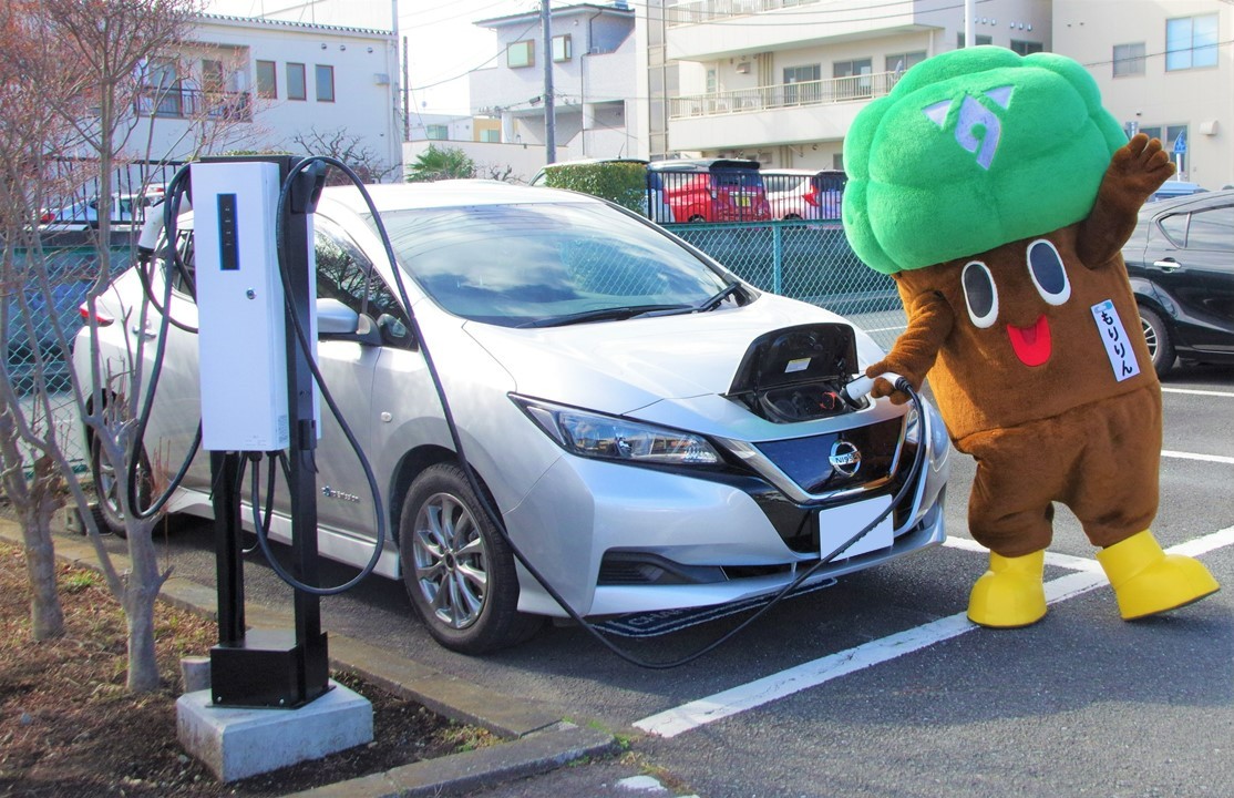 白い電気自動車が、屋外の駐車場に設置された充電器の前に停まり、フードを開けてマスコットキャラクター「もりりん」充電ケーブルを差し込んで充電している写真