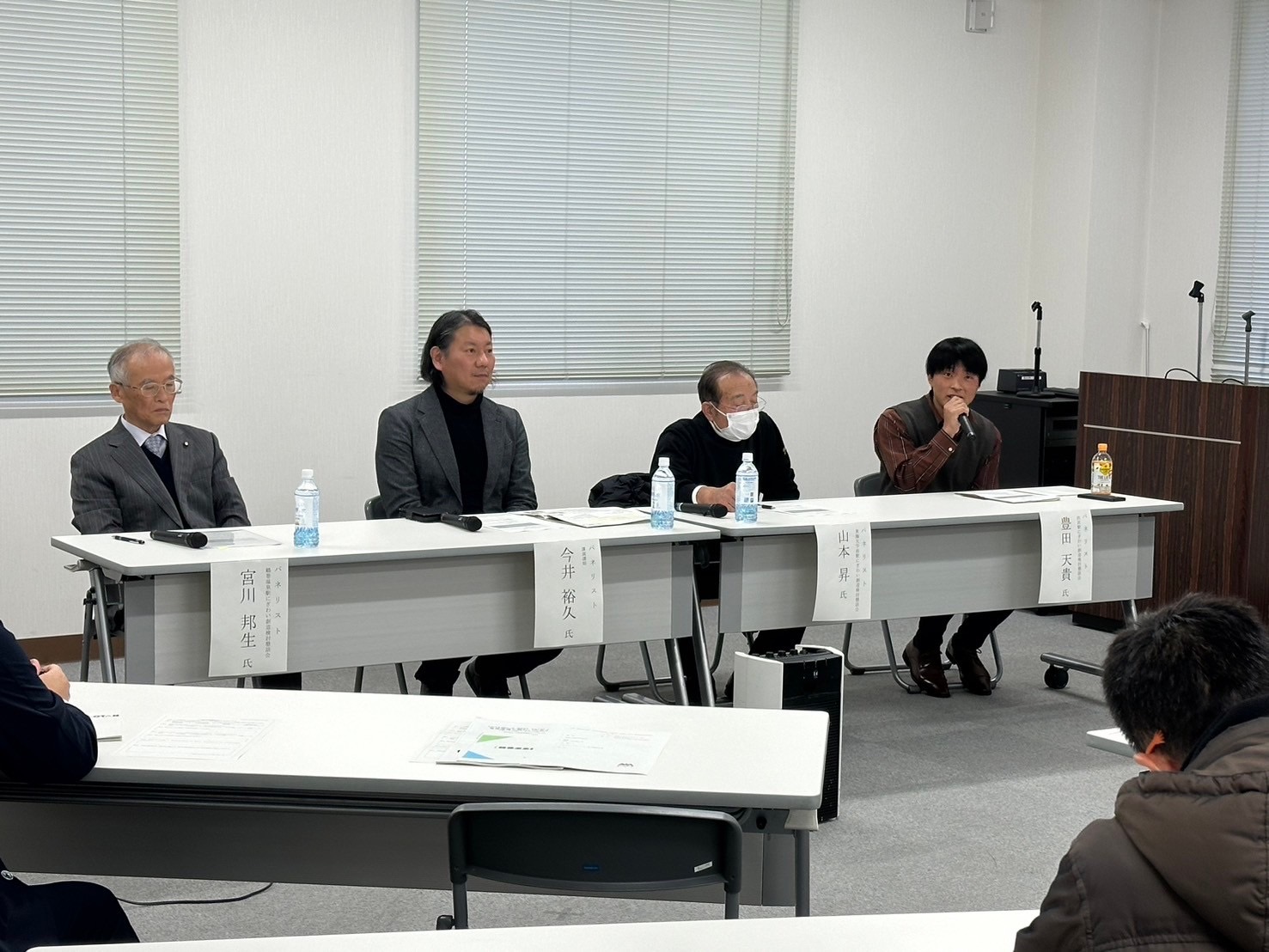 白色のブラインドがかかる窓を背景に、4人の男性が「宮川弥生氏」「金井啓一」「山崎淳」「豊田大賀」と記された登壇者札が置かれた長机に座り、マイクを前にしてパネルディスカッションを行っている風景写
