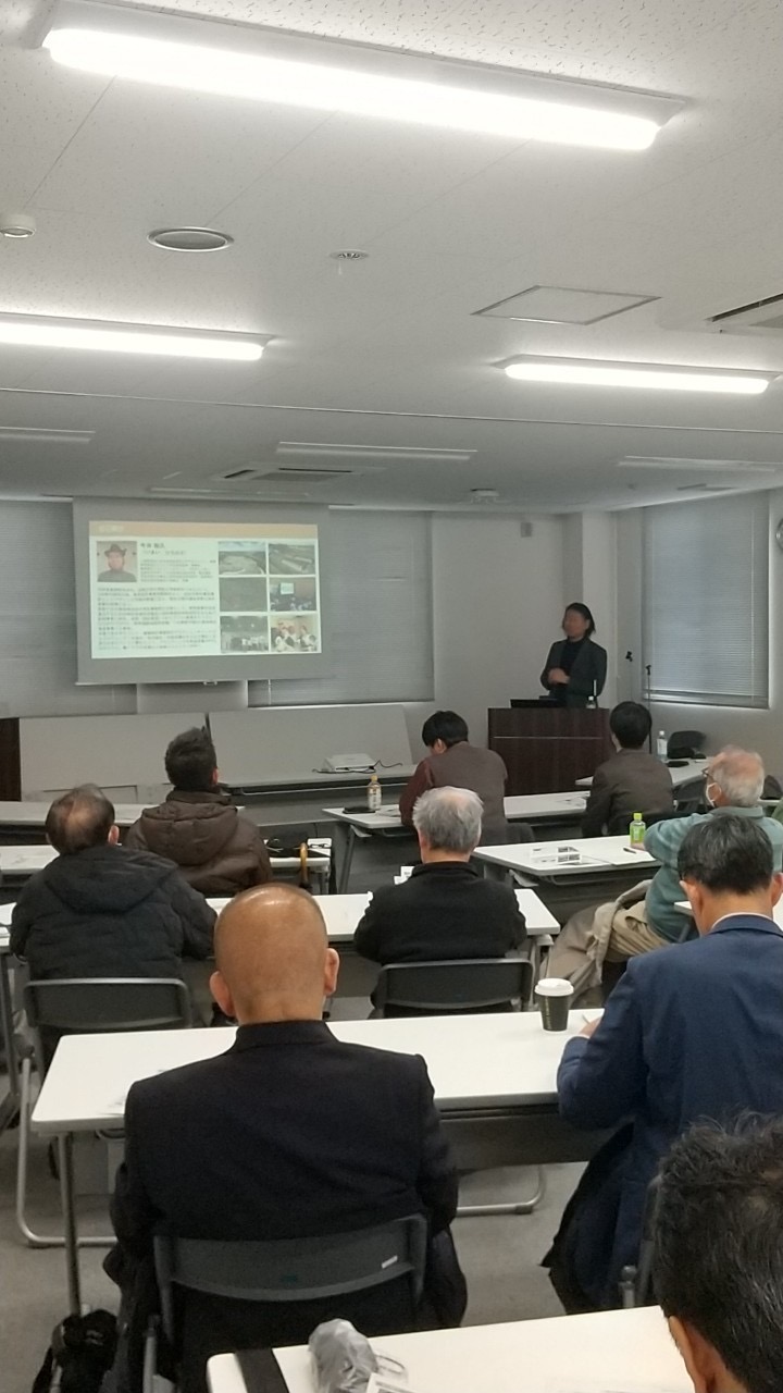 背中を向けて座る中高年の参加者たちの前で、講師の方がプロジェクターのスクリーンに映し出された資料を背景に、演台に立ち話している様子を後方から捉えた写真