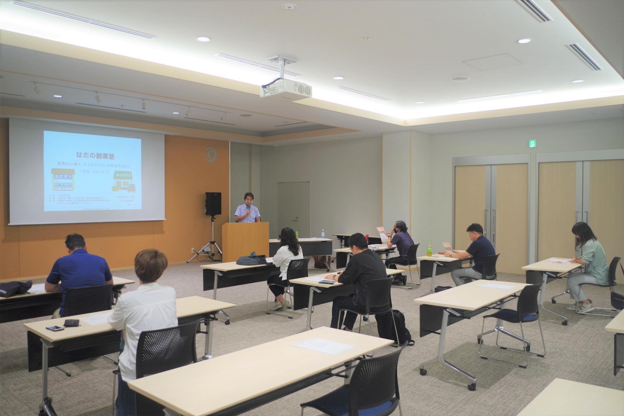 明るい照明の会議室で、演台に立つ白いシャツの講師が左側のスクリーンに「はだの創業塾」のスライドを投影し、前方に配置された机に着席する数名の受講者が講義を受けている様子の写真