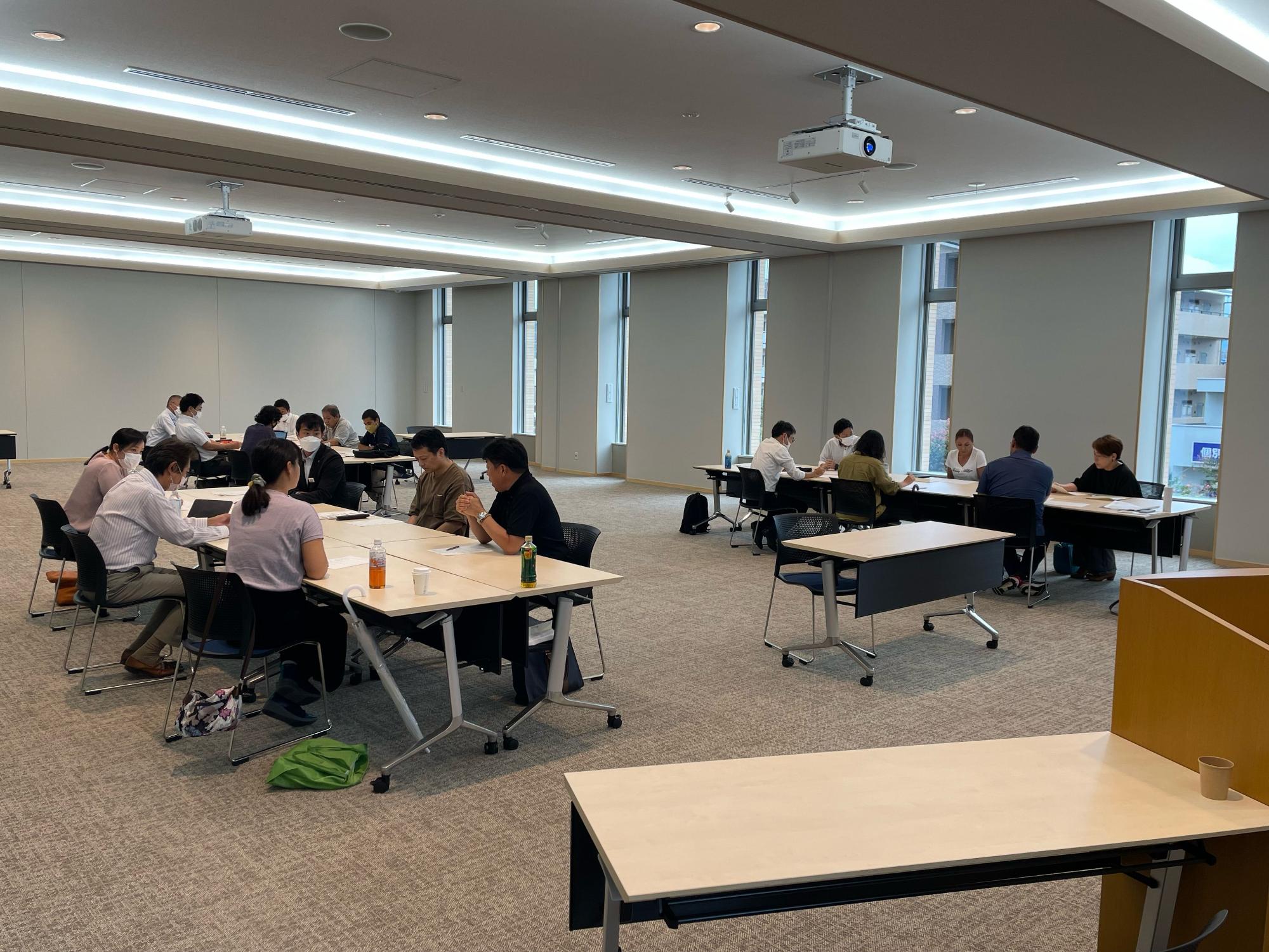 間接照明とプロジェクター、大きな窓のある明るい会議室で、参加者がマスクを着用し複数のテーブルに分かれて着席しグループワークを行っている写真