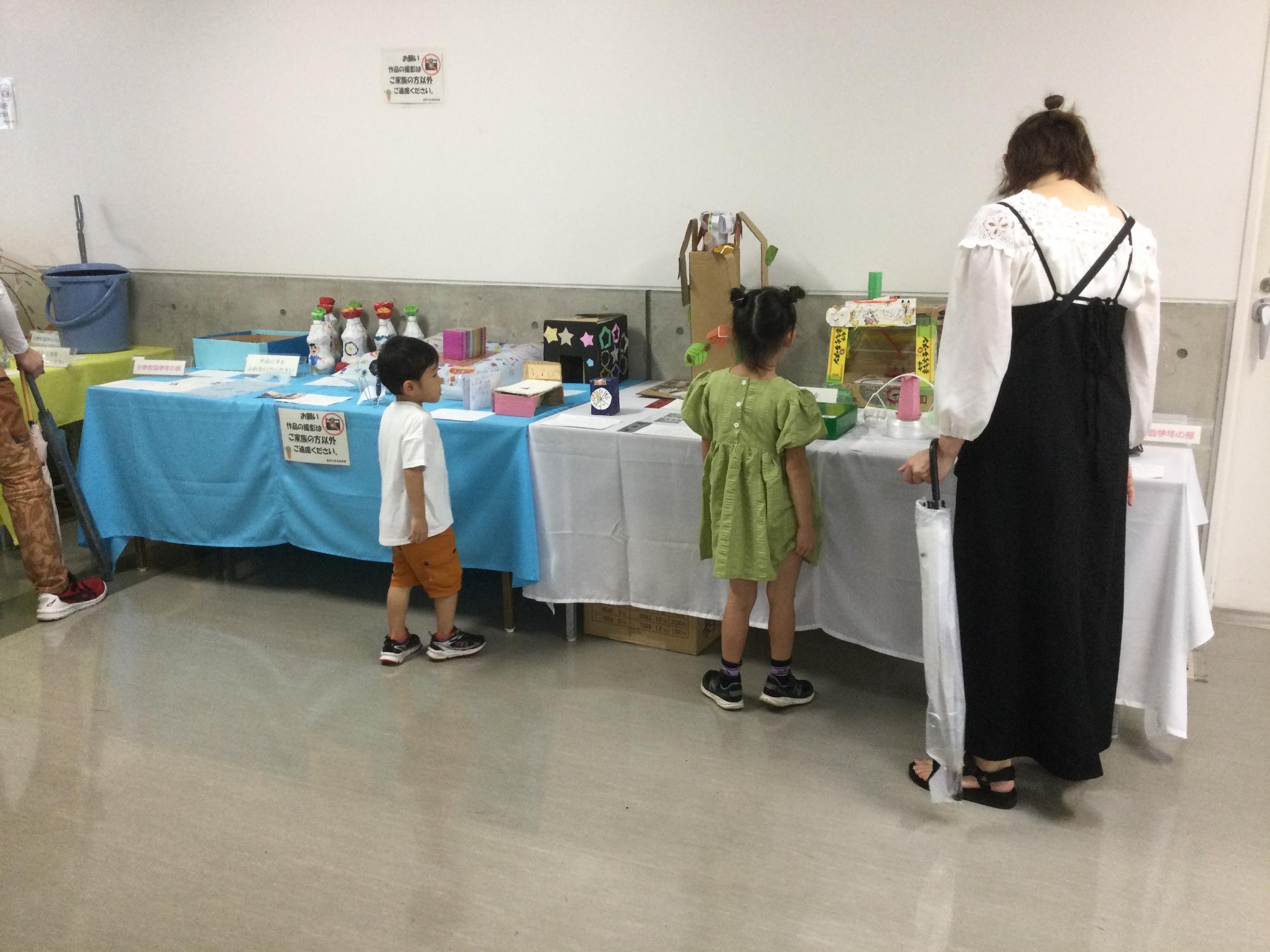 白い壁の室内で、2人の子供と母親の家族連れが水色や白色のテーブルクロスがかけられた創意工夫展の長机の上にに並べられた子どもたちの工作や発明品を鑑賞している様子の写真