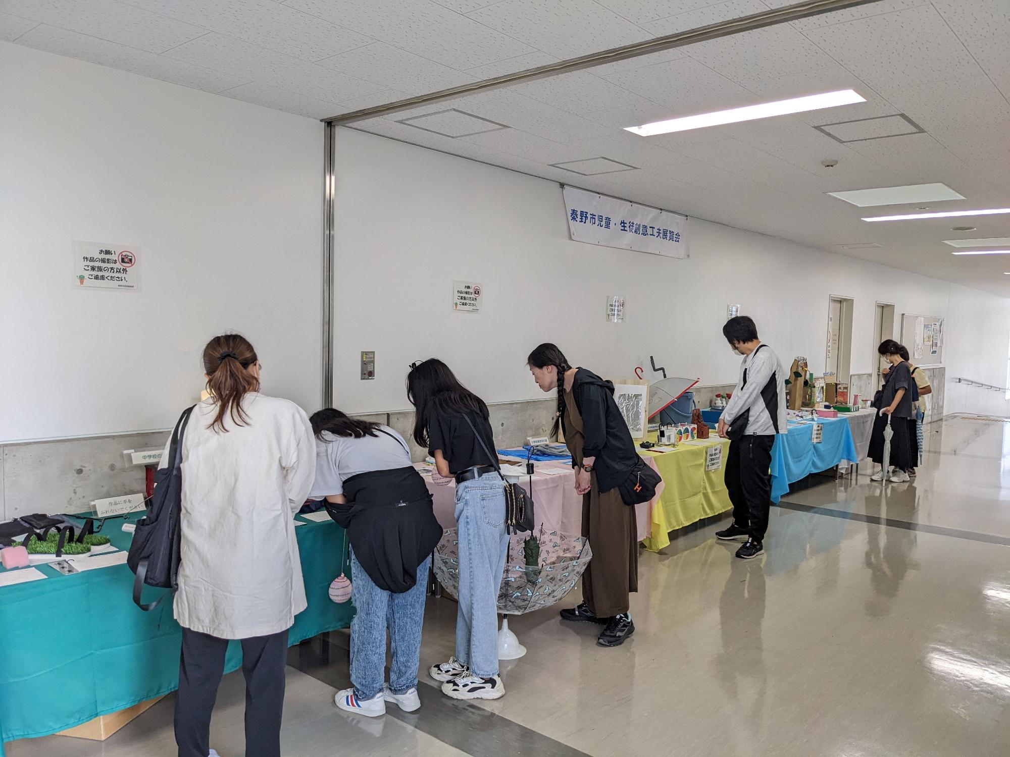 「秦野市児童・生徒創意工夫展覧会」と書かれた幕が掲示された白い壁沿いに、カラフルな色のテーブルクロスがかけられた長机が設置され、テーブルの上に並んだ子供たちの作品を、数人の来場者が興味深そうに覗き込んでいる、創意工夫展の様子を撮影した写真
