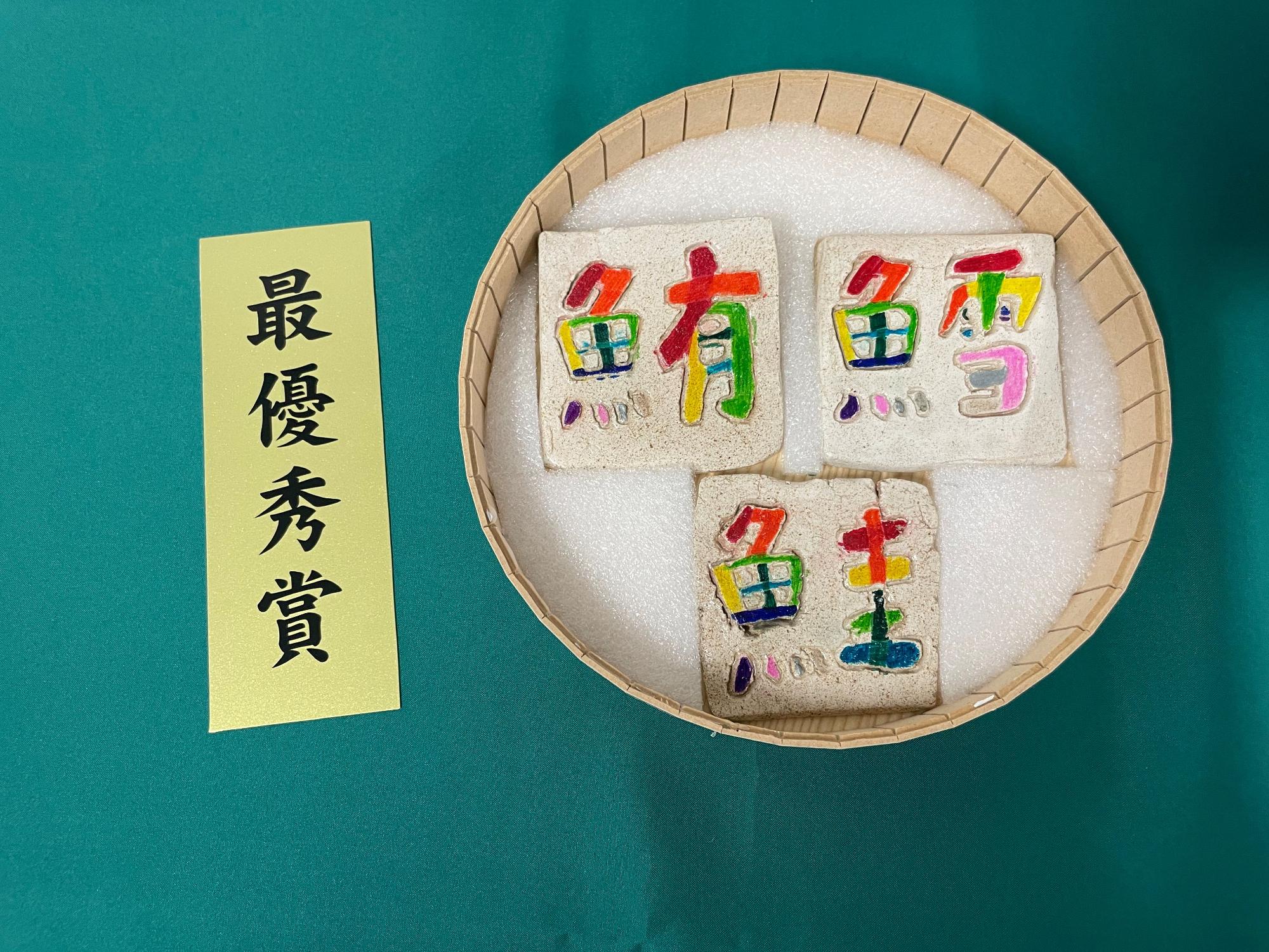 左側には「最優秀賞」と書かれた黄色い短冊が置かれており、白い発泡スチロール材が間に敷かれた円形の竹製の桶に、「鮪」、「鱈」、「鮭」という漢字が彫刻されており、カラフルなクレヨンやペンキで着色されている3つの正方形の石のプレートの作品が並べられている写真