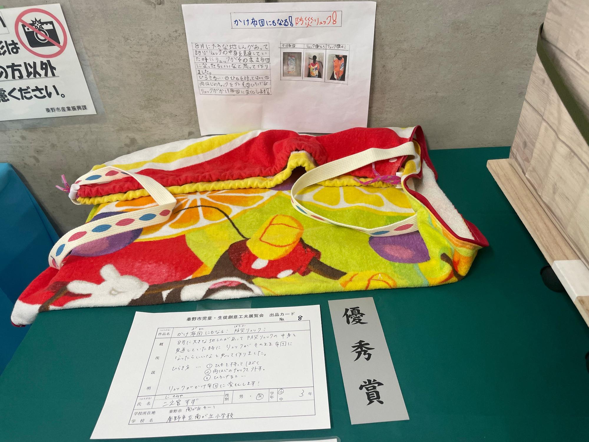 右側に「優秀賞」と書かれたグレー色の短冊が置かれており、使い方を説明する写真や図が壁に貼られ、両端に幅の太い白いゴムのような物が縫われたミッキーマウス柄のカラフルなタオル地の作作品が展示されている写真