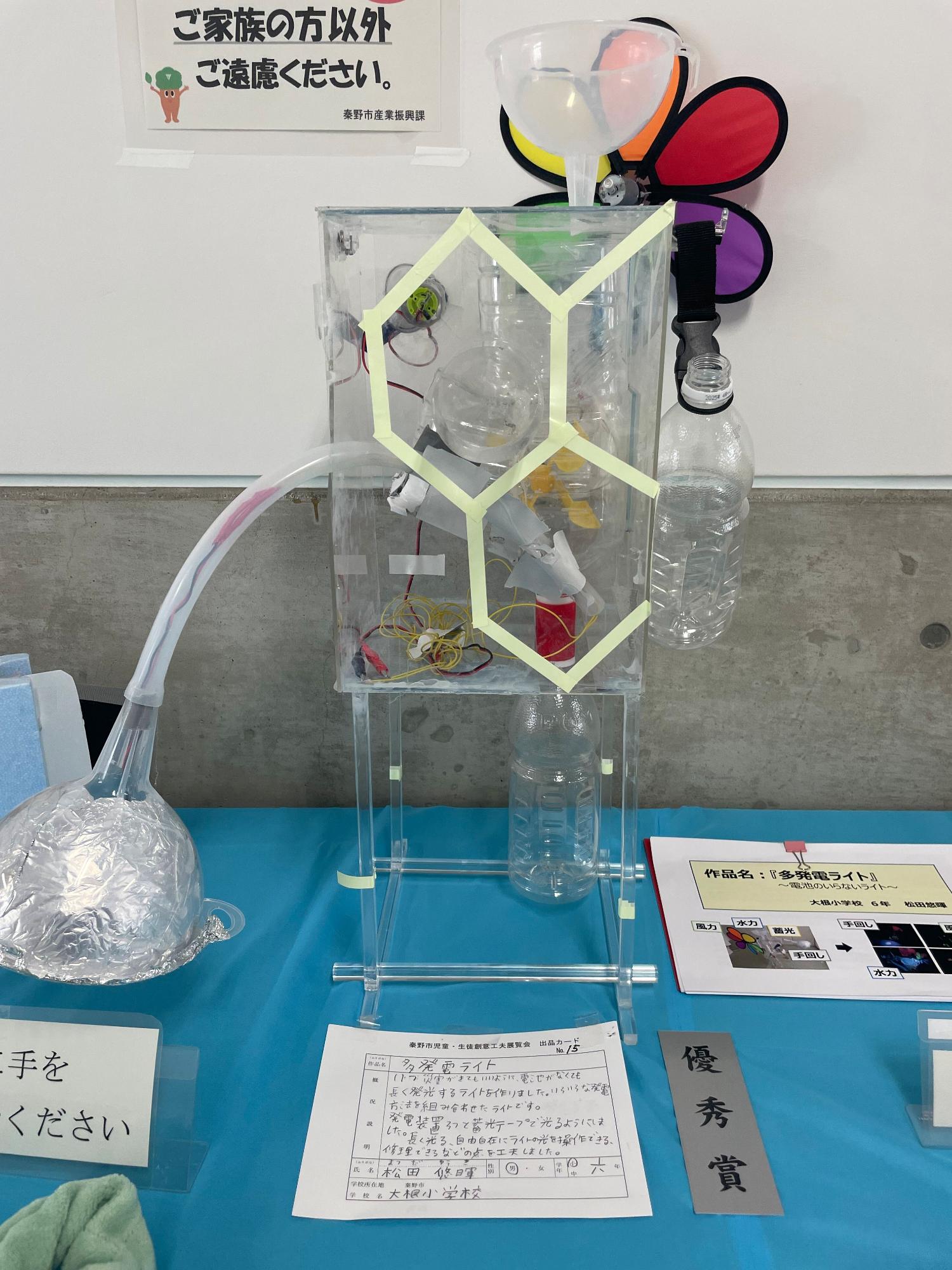 「優秀賞」と書かれたグレー色の短冊と出品カードが置かれており、アクリル板でできた透明な箱に、ペットボトルや導線、ビニールホースなどが組み込まれ、その周りには蛍光色のテープでハチの巣のような六角形の模様が付けられている、小学生が製作した「多発電ライト」という作品の写真
