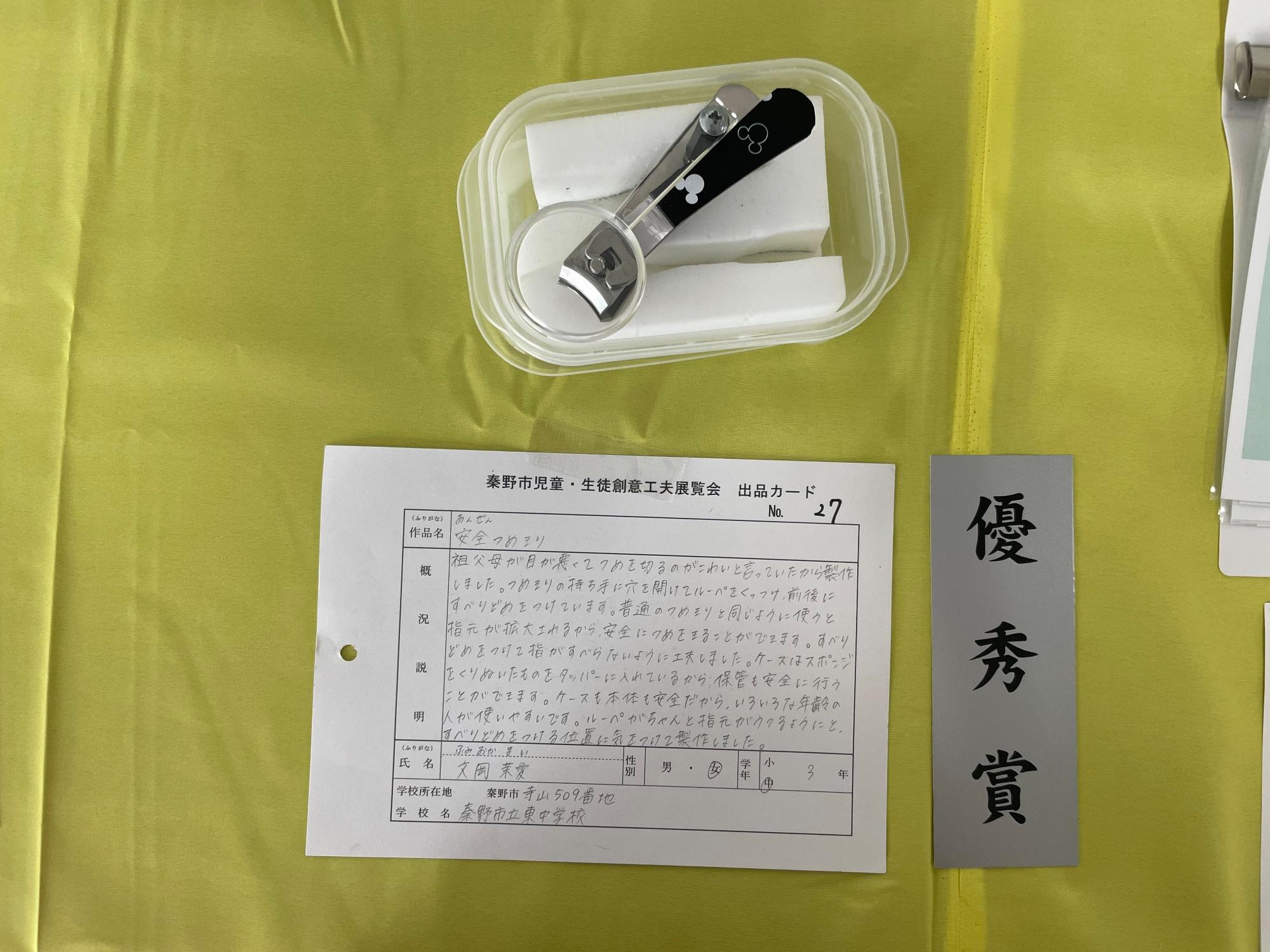 「優秀賞」と書かれたグレー色の短冊と出品カードが置かれており、カットされた白いスポンジが敷かれた透明のプラスチック容器に、ルーペを取り付けたミッキーマウス柄の黒い爪切りの作品の写真