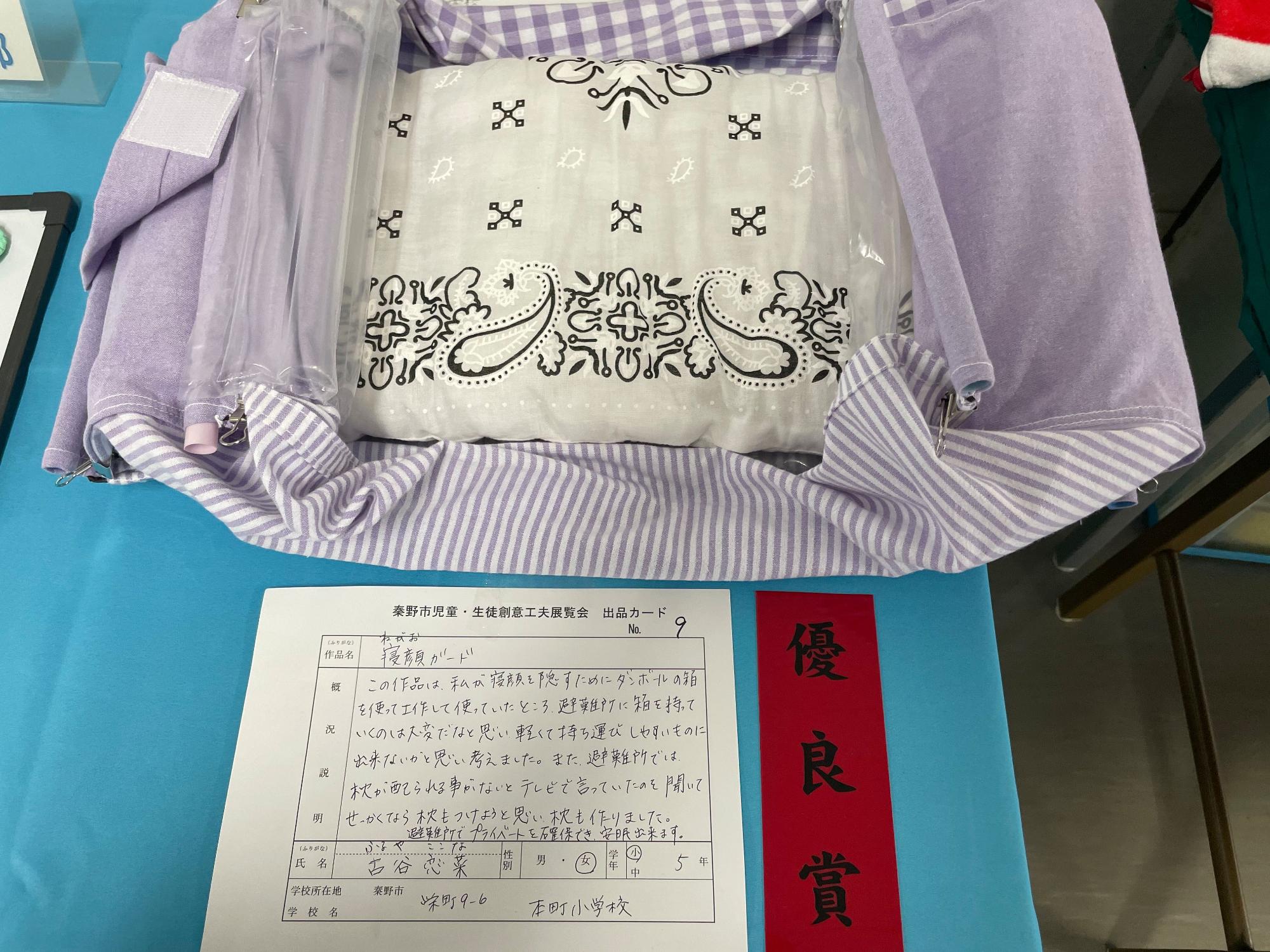 「優良賞」と書かれた赤色の短冊と出品カードが置かれており、左側に正方形の粘着テープが取り付けられ、四方がクリップで留められた手製の紫のギンガムチェック柄の布と、その中にしまわれている黒いペイズリー柄のバンダナで作られた枕が展示されている「寝顔ガード」というタイトルの作品の写真