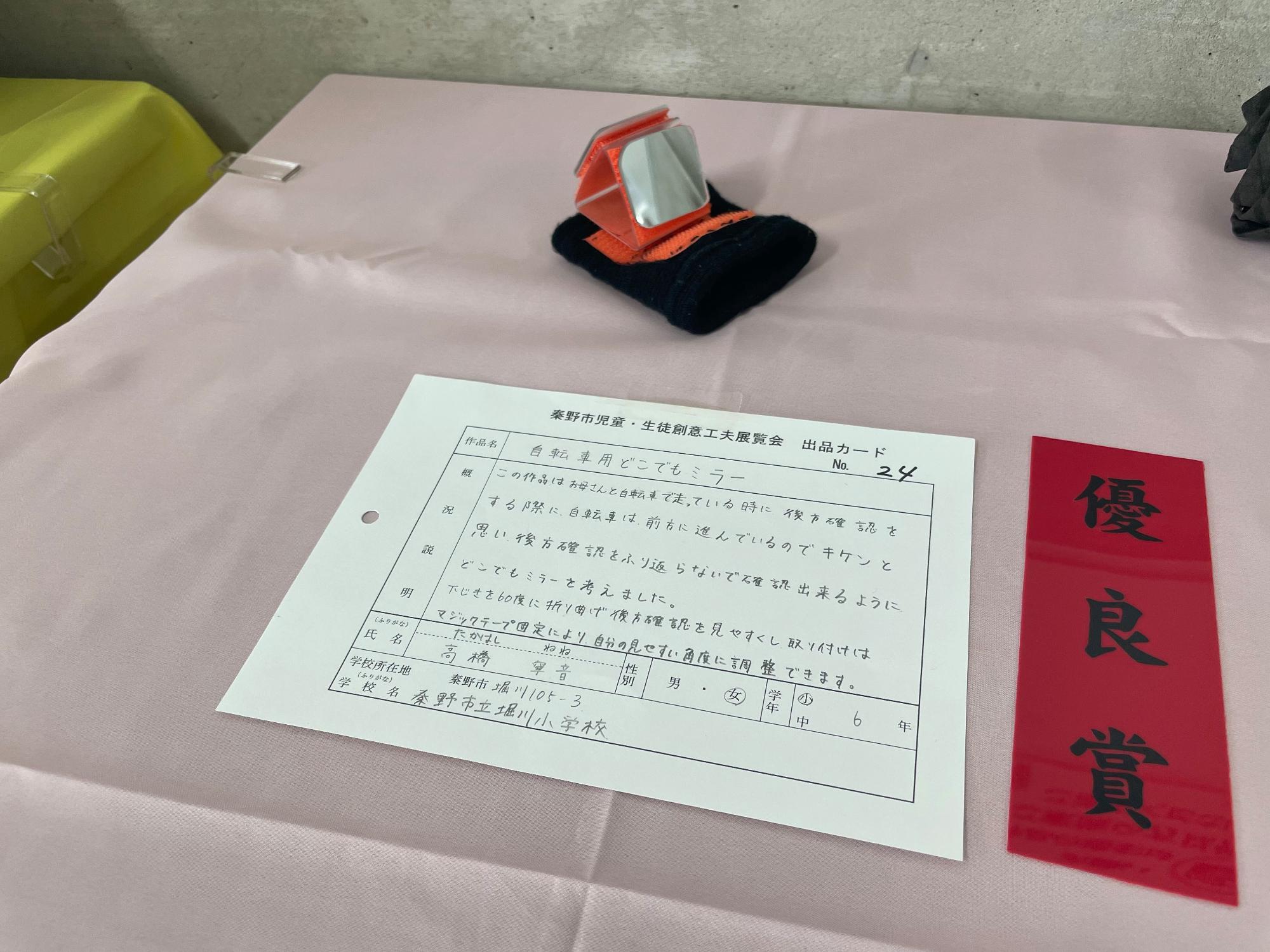 「優良賞」と書かれた赤色の短冊と出品カードが置かれており、オレンジ色のマジックテープが縫われた黒いリストバンドの上に、四角形のミラーが三角形に折り曲げたプラスチック板の前後に取り付けられた「自転車用どこでもミラー」というタイトルの作品の写真