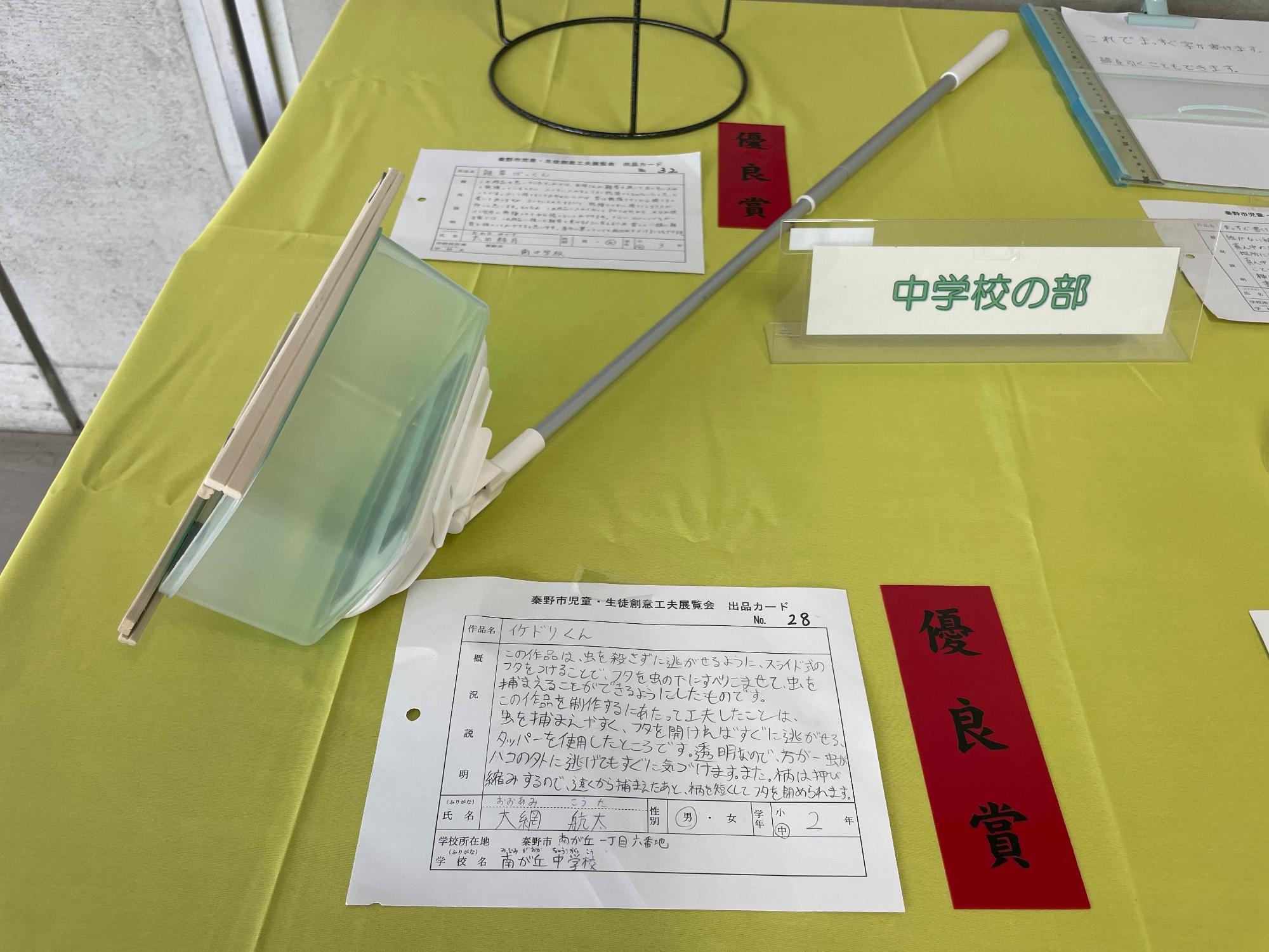 「優良賞」と書かれた赤色の短冊と出品カードが置かれており、プラスチック製の容器が取り付けられた伸び縮みする柄の作品が展示された「イケドリくん」というタイトルの作品の写真