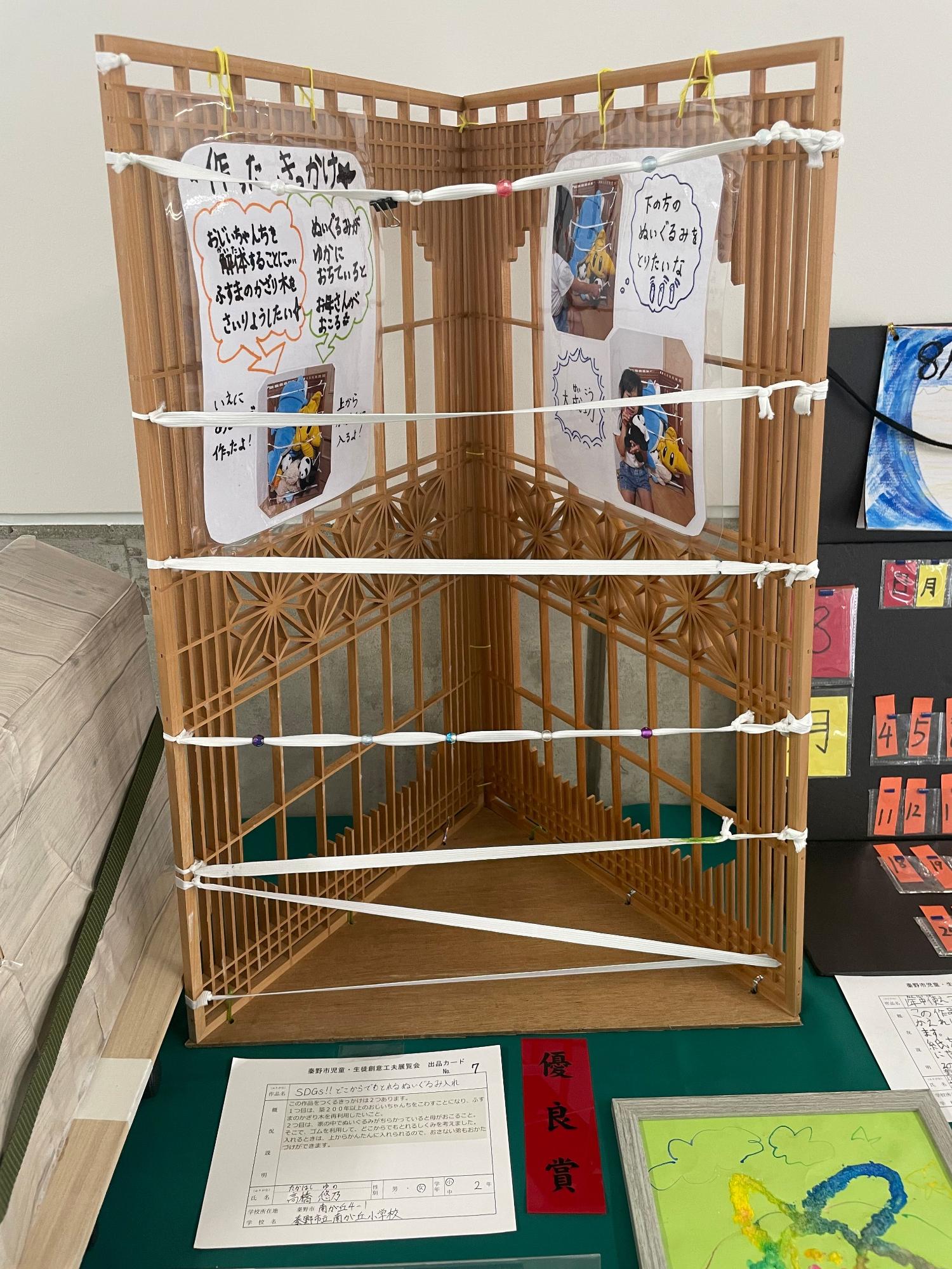 「優良賞」と書かれた赤色の短冊と出品カードが置かれており、三角形の木製の土台の上に2つの木製のかざり木を取り付け、表面に等間隔で白いゴムを取り付けた、作ったきっかけなどが手書きで記載され写真が添えられた用紙と共に展示された、「SDGs!!どこからでもとれるぬいぐるみ入れ」というタイトルの作品の写真