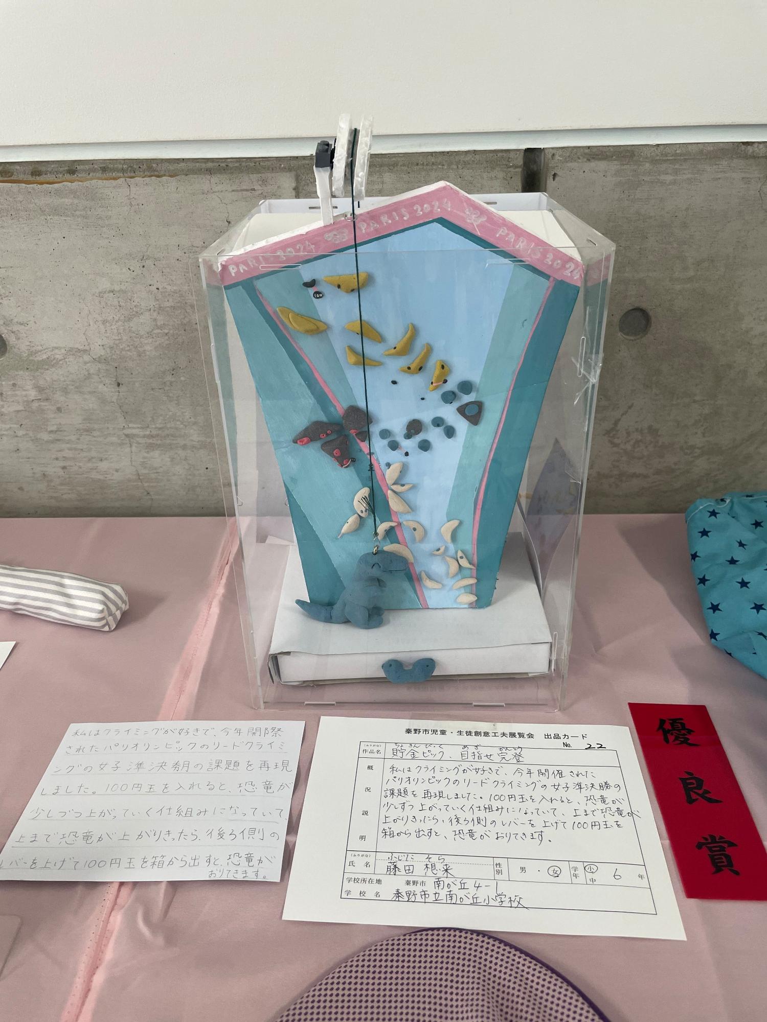 「優良賞」と書かれた赤色の短冊と出品カードが置かれており、長方形の透明ケースの中に、様々な形や色をした貝殻や石が水色や青色で塗られた立体的な岩壁登りのコースに付けられ、壁の頂上から恐竜のキャラクターがぶら下がっている「貯金ピック、目指せ完登」というタイトルの作品の写真