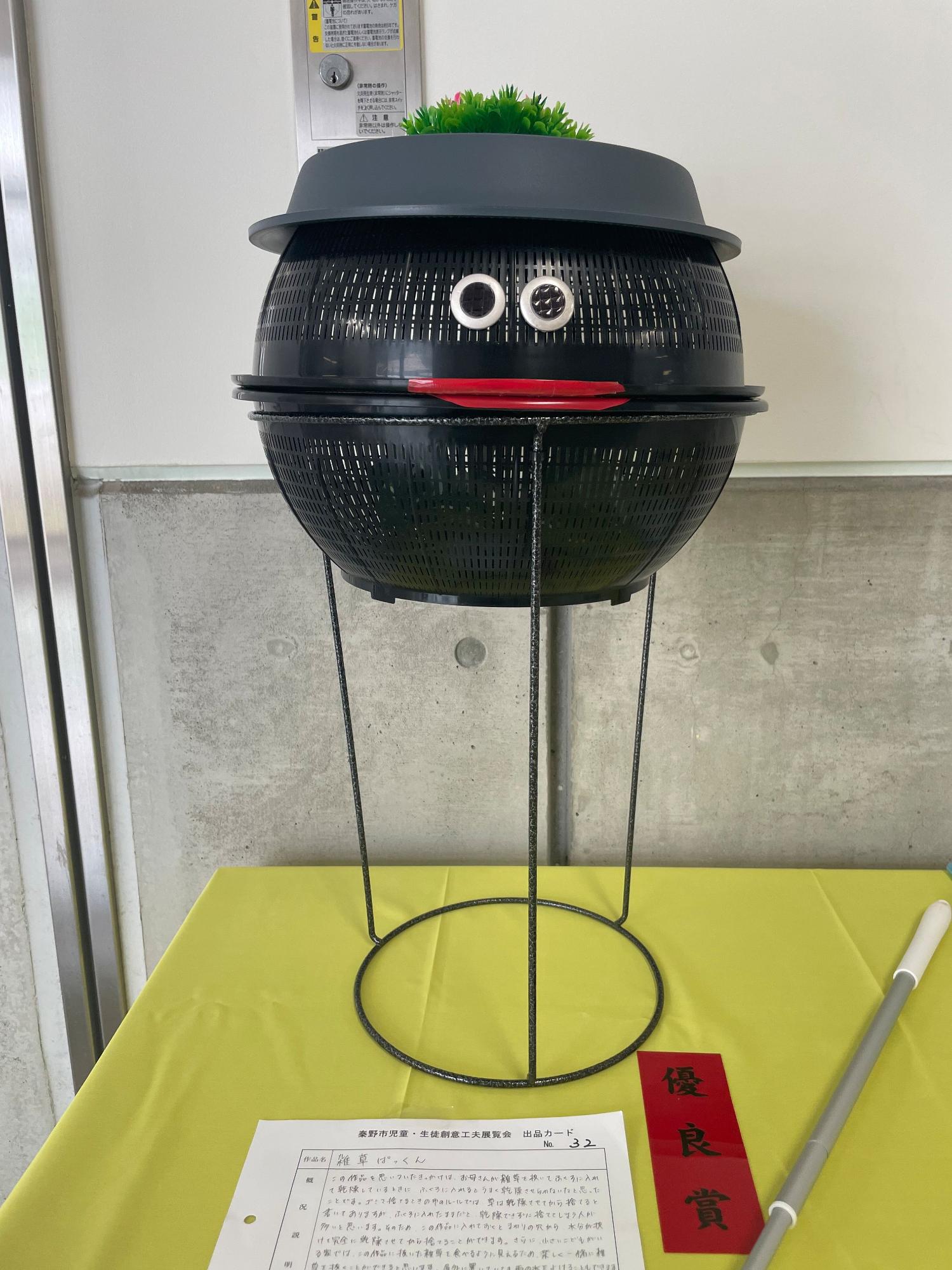 「優良賞」と書かれた赤色の短冊と出品カードが置かれており、黒い網を上下に2つ重ね、黒い水受け皿を逆さまに置き帽子をかぶった顔のような形に作り、そこに目や赤い口がつけられた作品がポールプランタースタンドに置かれている「雑草ぱっくん」というタイトルの作品の写真