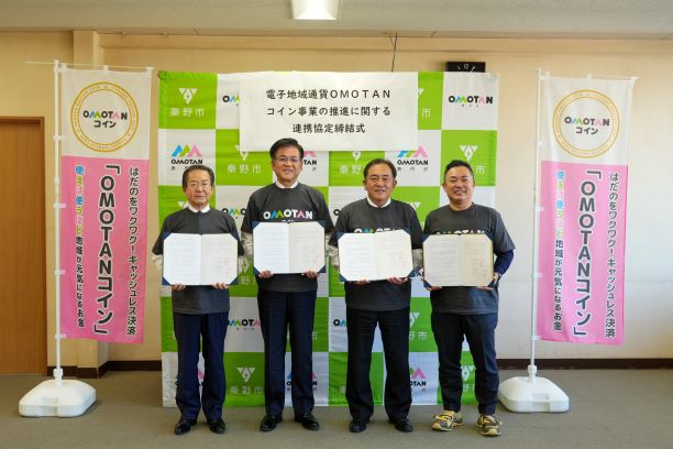 「OMOTANコイン」ののぼり旗が並ぶ前で、四人の男性が各々協定書らしき書類を開いて持ち、カメラに向かって並んで立っており、中央の横断幕には「電子地域通貨 OMOTAN コイン事業の推進に関する連携協定締結式」と書かれている記念写真