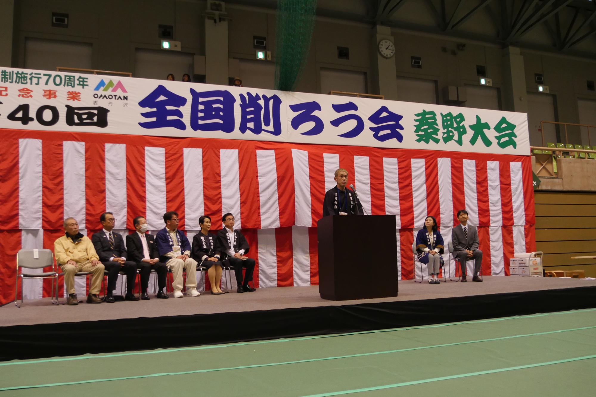 体育館のような会場の壇上に、白と赤の紅白幕を背景に「全国削ろう会 秦野大会」と書かれた横断幕が掲げられた式典で、中央の演台に立つ男性がスピーチを行い、その左右に複数人の関係者が着席している様子の写真