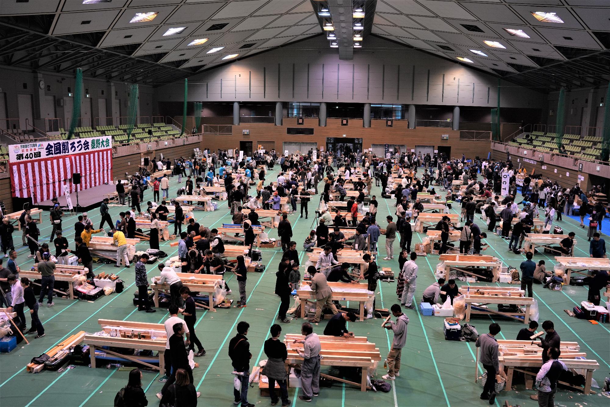 大きな体育館のような屋内施設で、多数の参加者が木工作業台を囲んで鉋などの道具を使って木材を削る作業に集中しており、奥には紅白幕のステージが見える、熱気あふれるイベント会場の全景を写した写真
