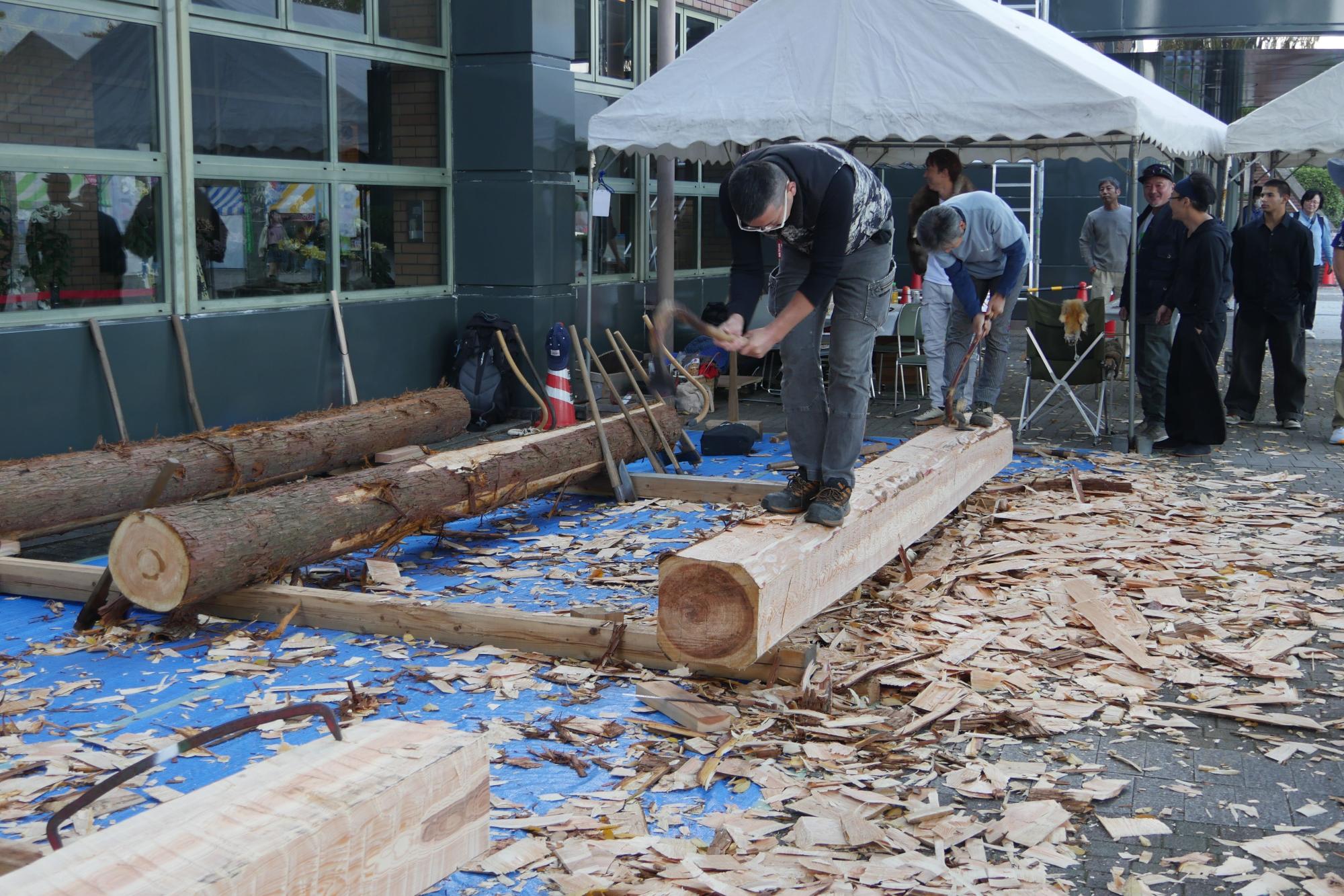 屋外の広場に敷かれた青いシートの上で、土木作業服の男性が大きな丸太に斧を振り下ろして皮むきや木材の加工を行っており、周囲には大量の削り屑が散乱している作業風景の写真