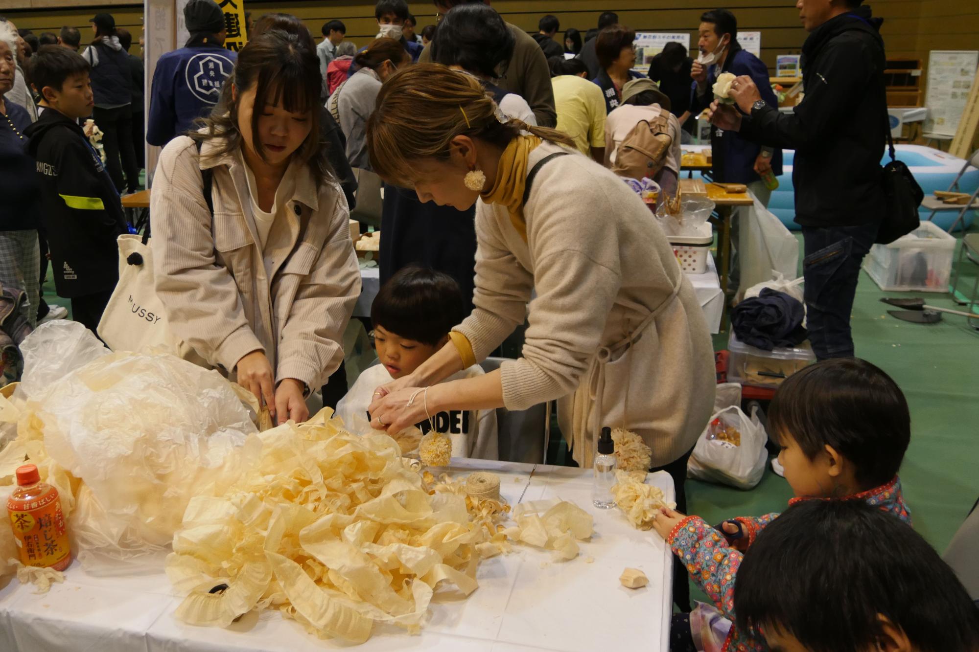 会場内のブースで、女性と子どもが、大量の薄い削り屑を使って、座っている小さな子どもに花のような飾りを作る体験を手伝っている様子の写真