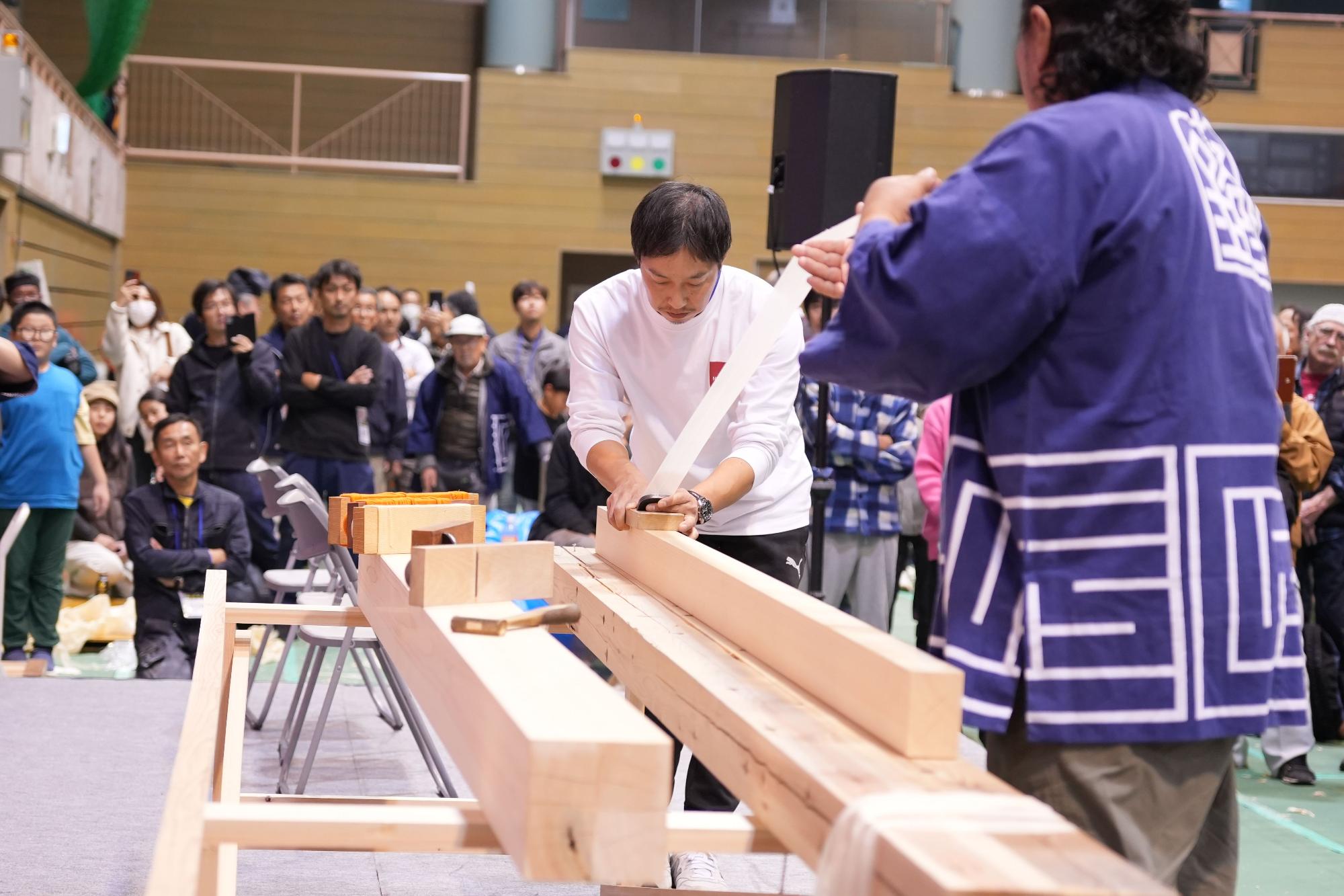 イベント会場で、白い長袖Tシャツを着た男性が、多数の観客に見守られながら、長く水平に置かれた木材の表面を、鉋を使って薄く削りだした木片を手に真剣な表情で実演している様子の写真