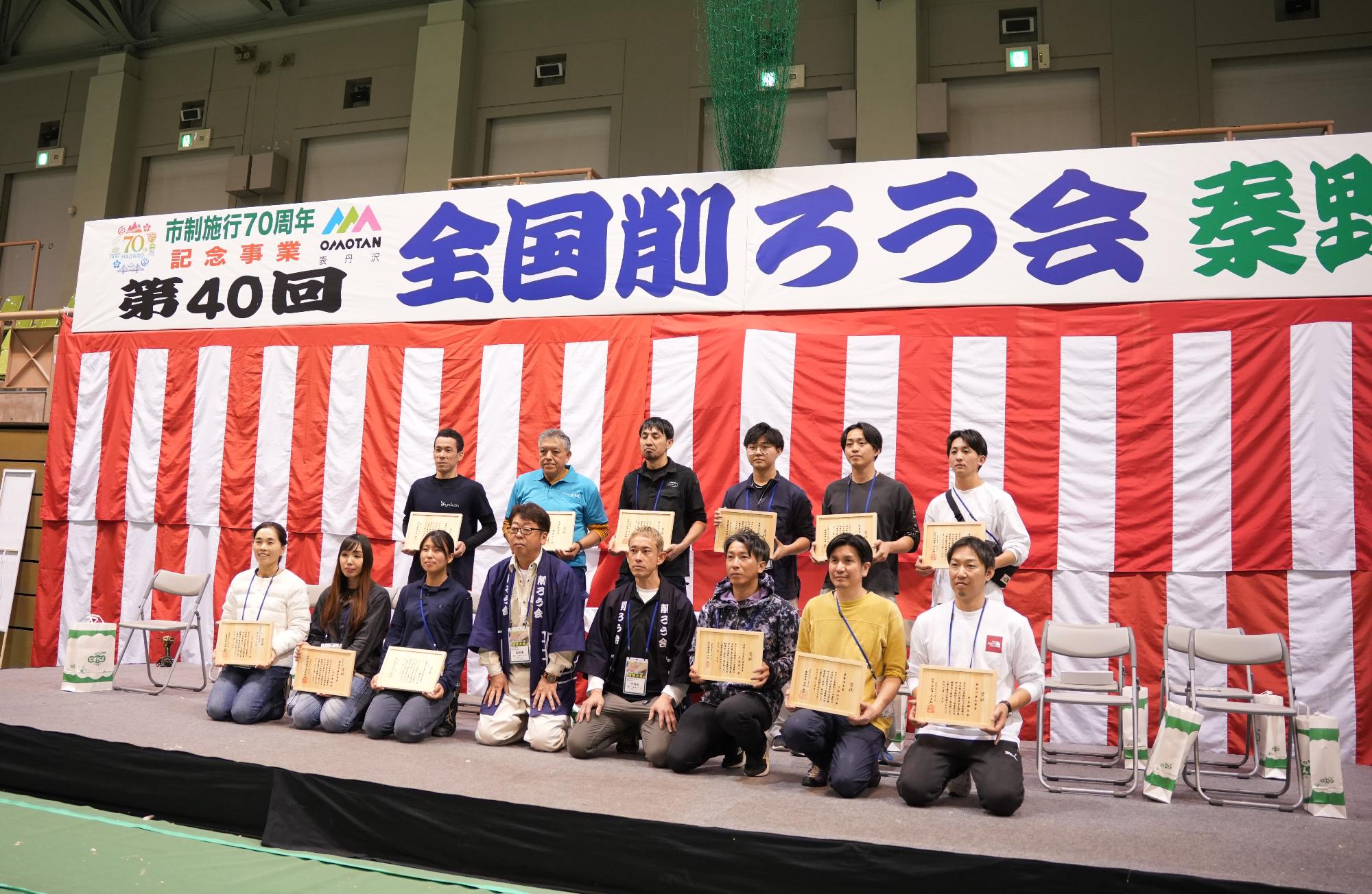 体育館内の壇上で、「第40回 全国削ろう会 秦野大会」の横断幕と紅白幕を背景に、競技で優秀な成績を収めたと思われる参加者の男女が賞状を手にし、笑顔で整列している集合写真